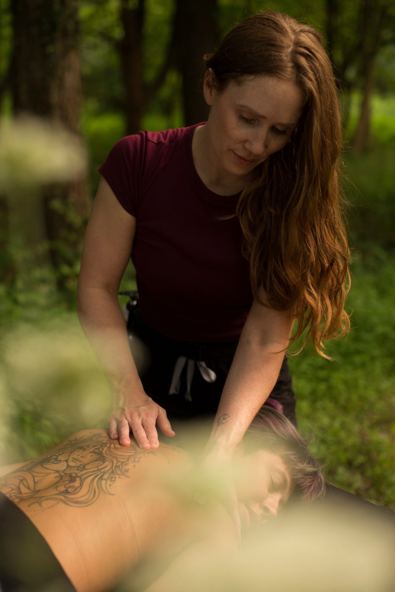Woman with long red hair leaning over a person with tattoos outdoors in a green park.