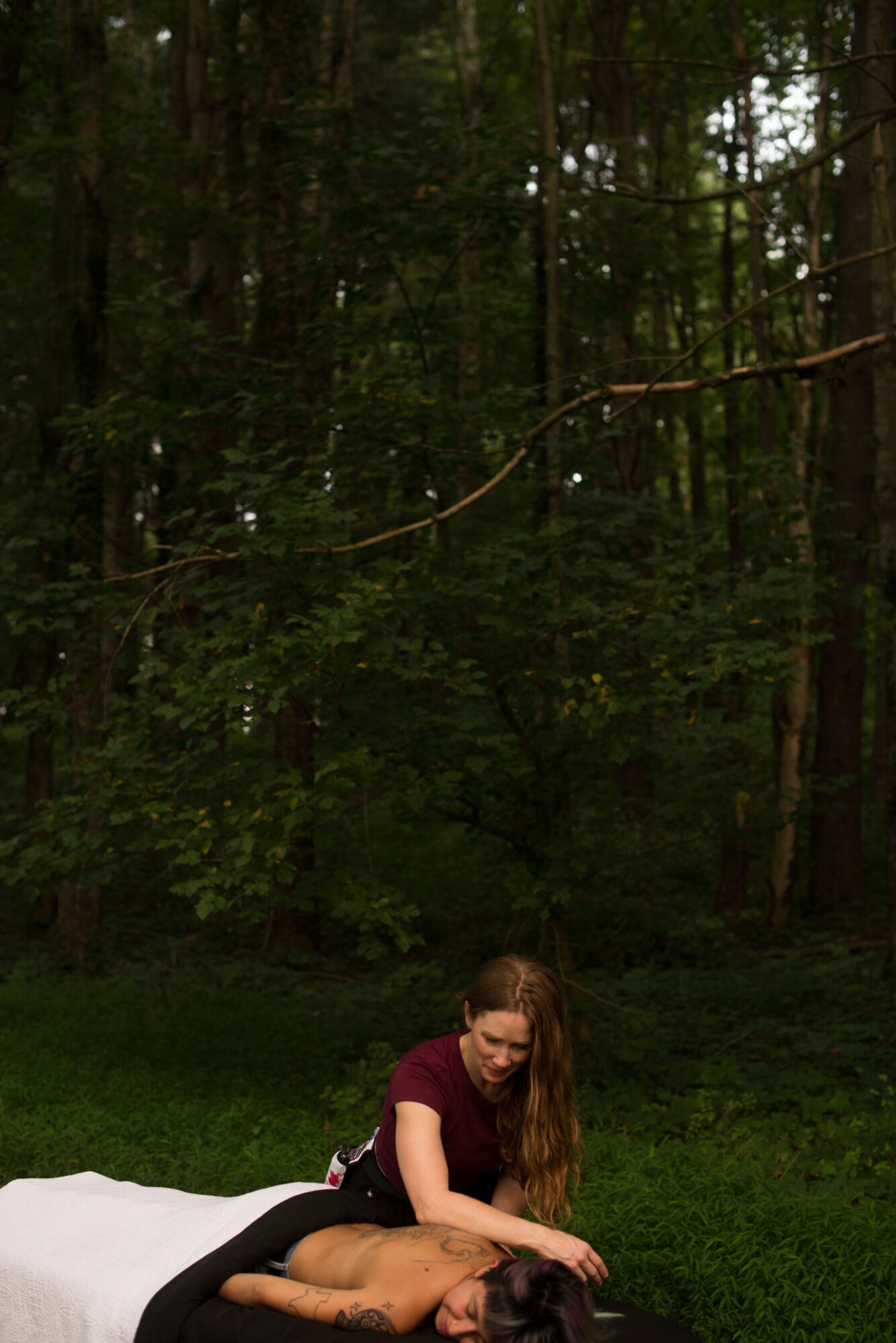 Person lying on a white surface outdoors with a woman kneeling beside them in a forest setting.