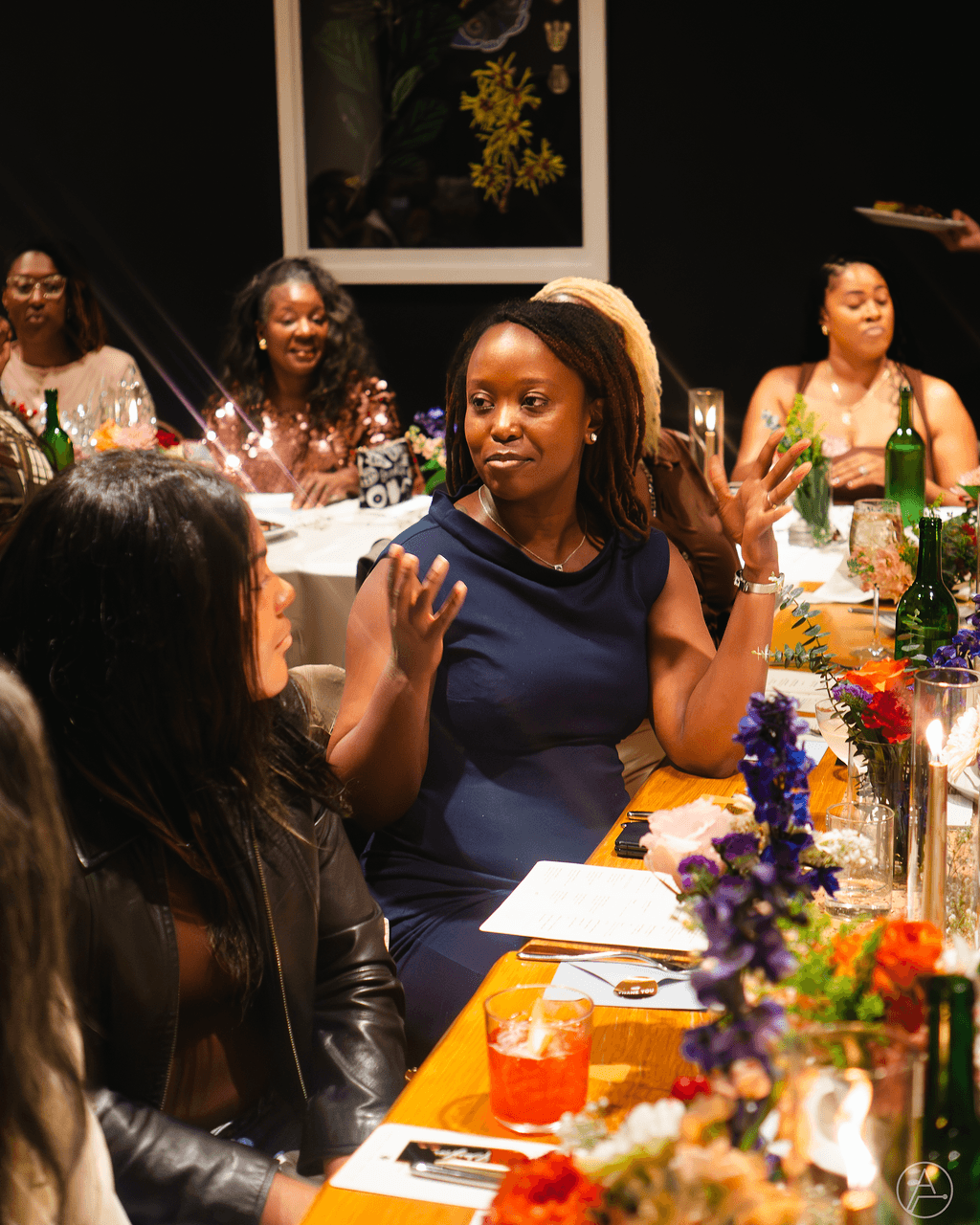 Group of women sitting at a decorated table, engaging in conversation, with drinks and flowers, in a dimly lit setting.