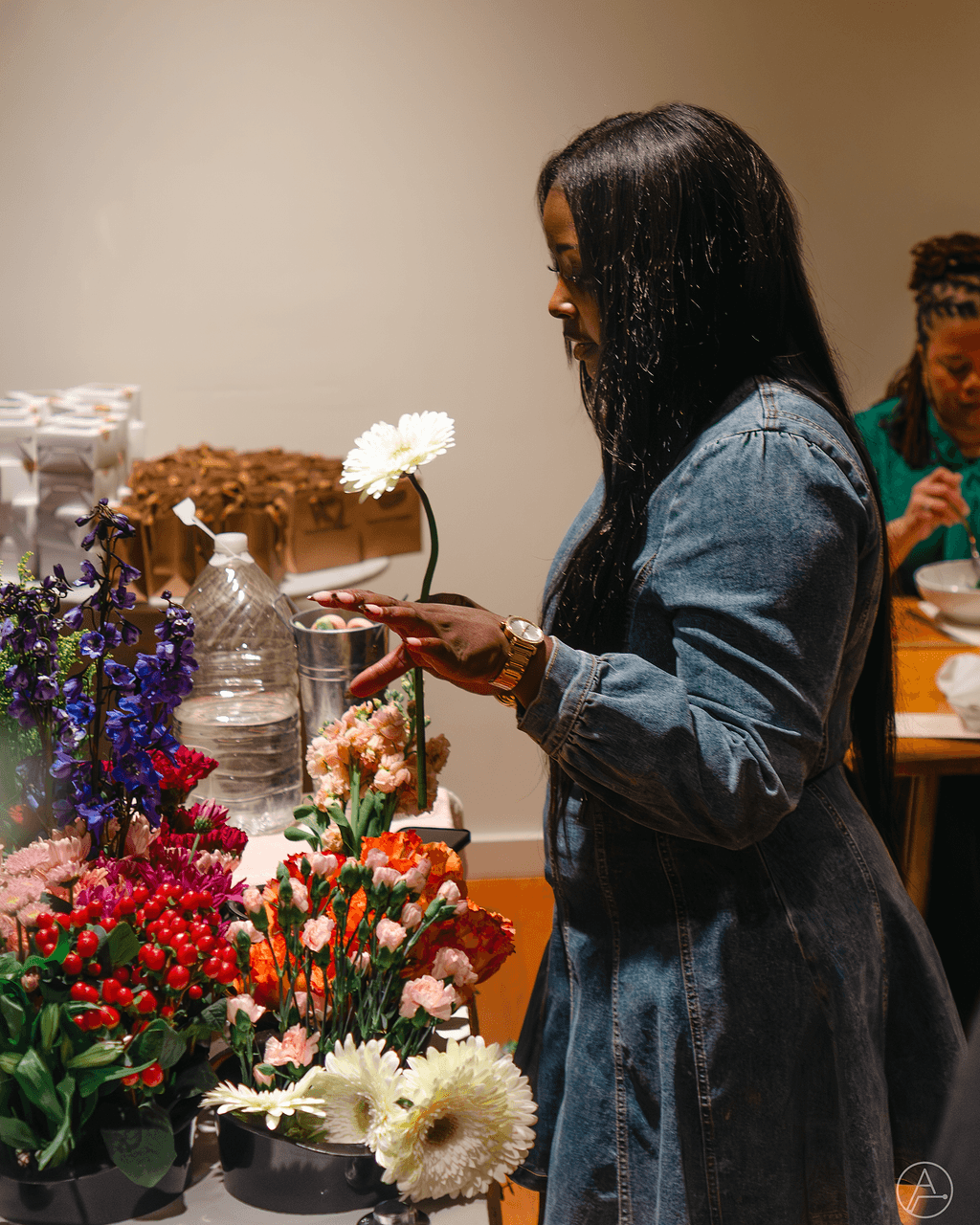Woman in denim jacket looking at her phone near colorful flowers and table with food, indoors.
