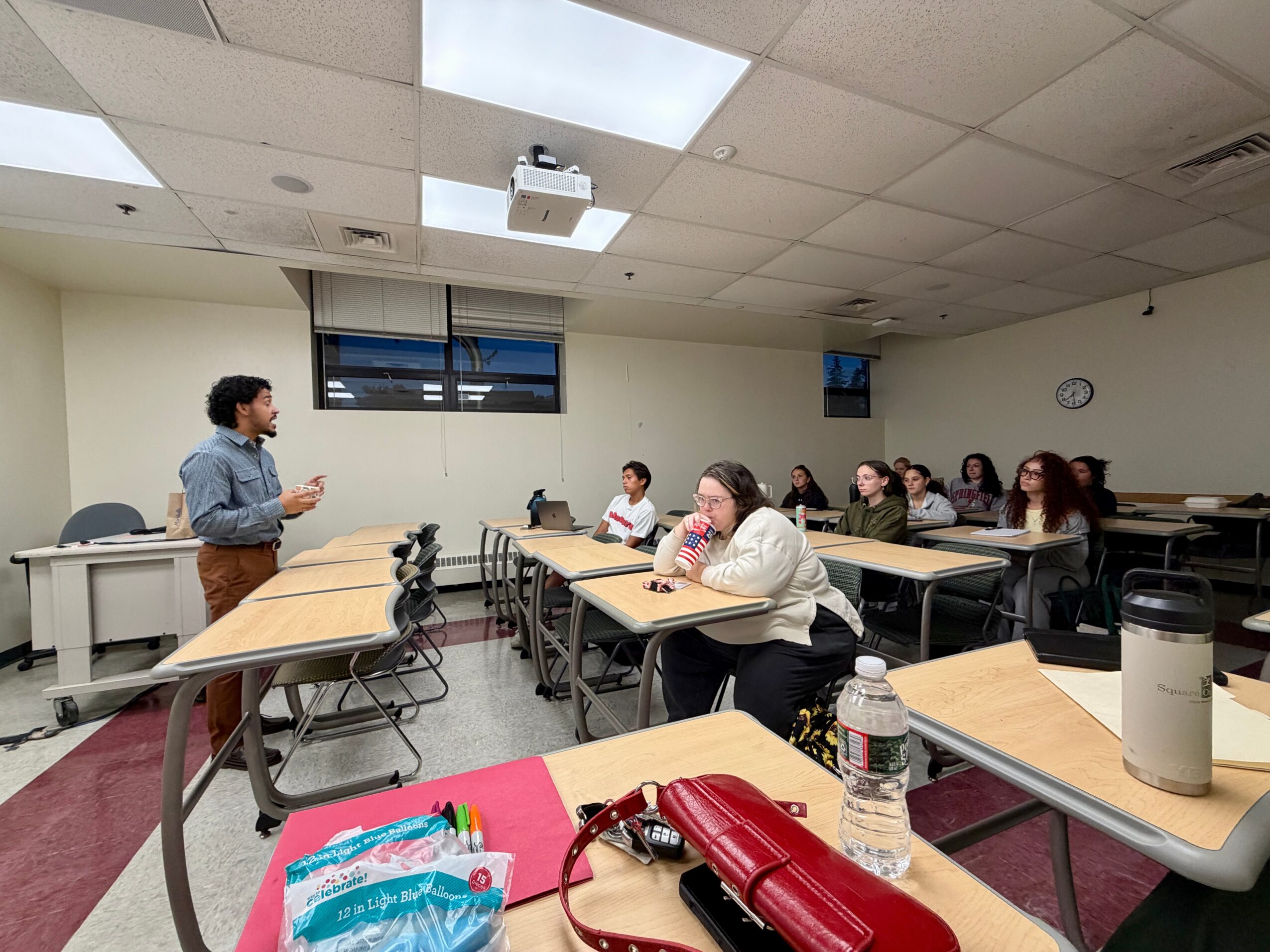 A person stands at the front of a classroom speaking to students seated at desks, with a projector on the ceiling.