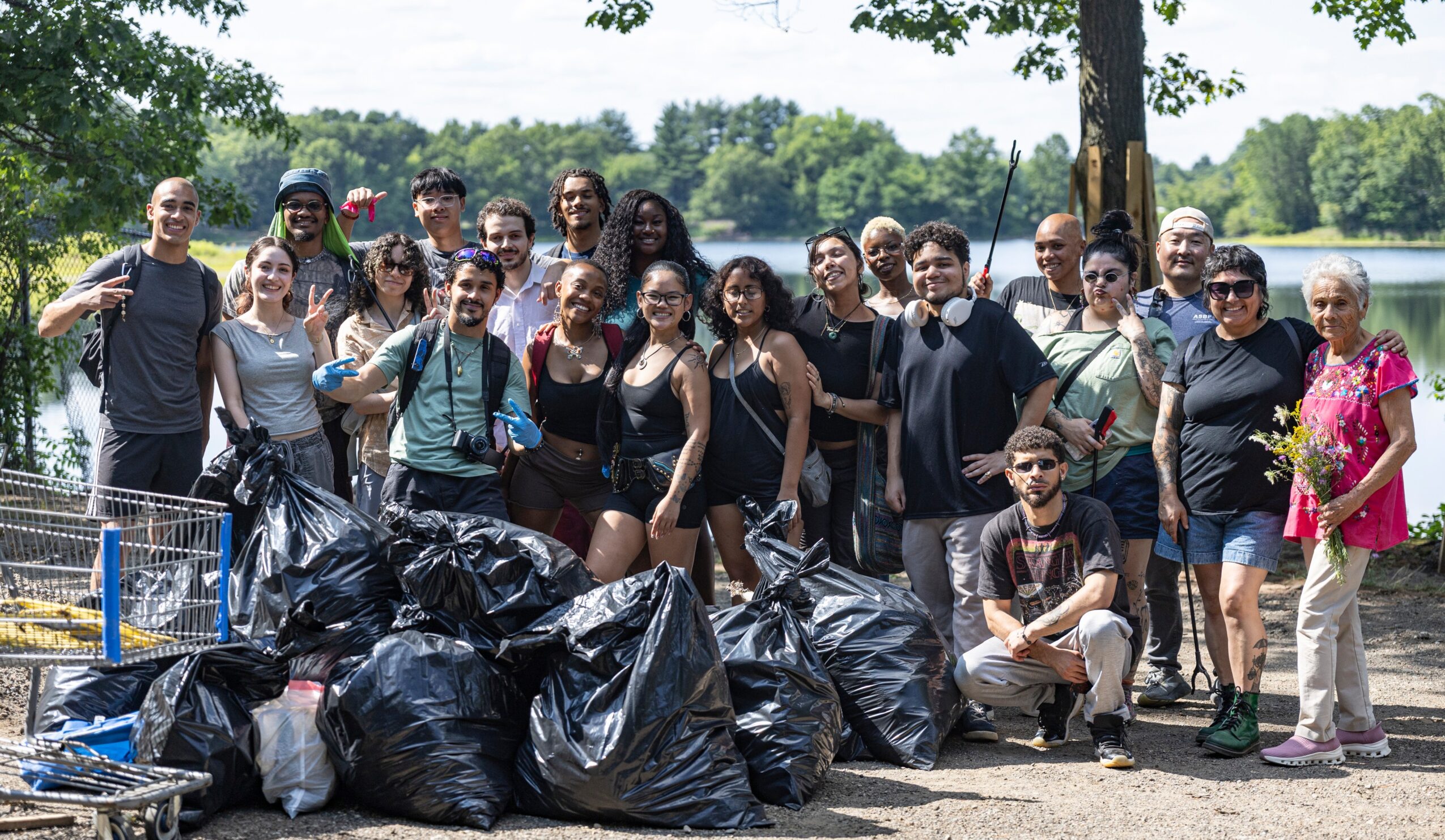 Group of people outdoors with trash bags, trees, and a clear sky in the background, participating in a cleanup event.