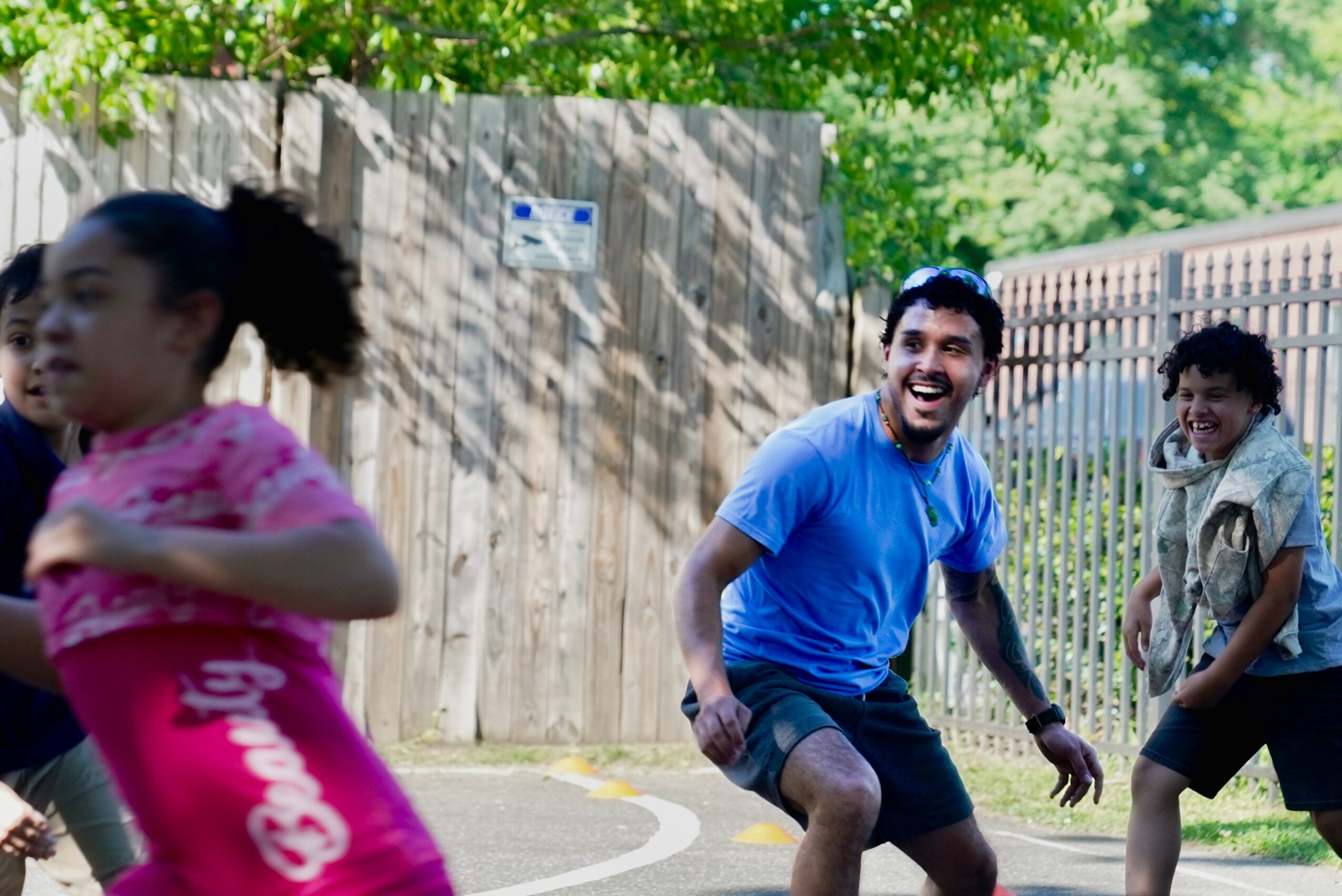 Three people outdoors near a wooden fence, smiling and playing, with trees and sunlight in the background.