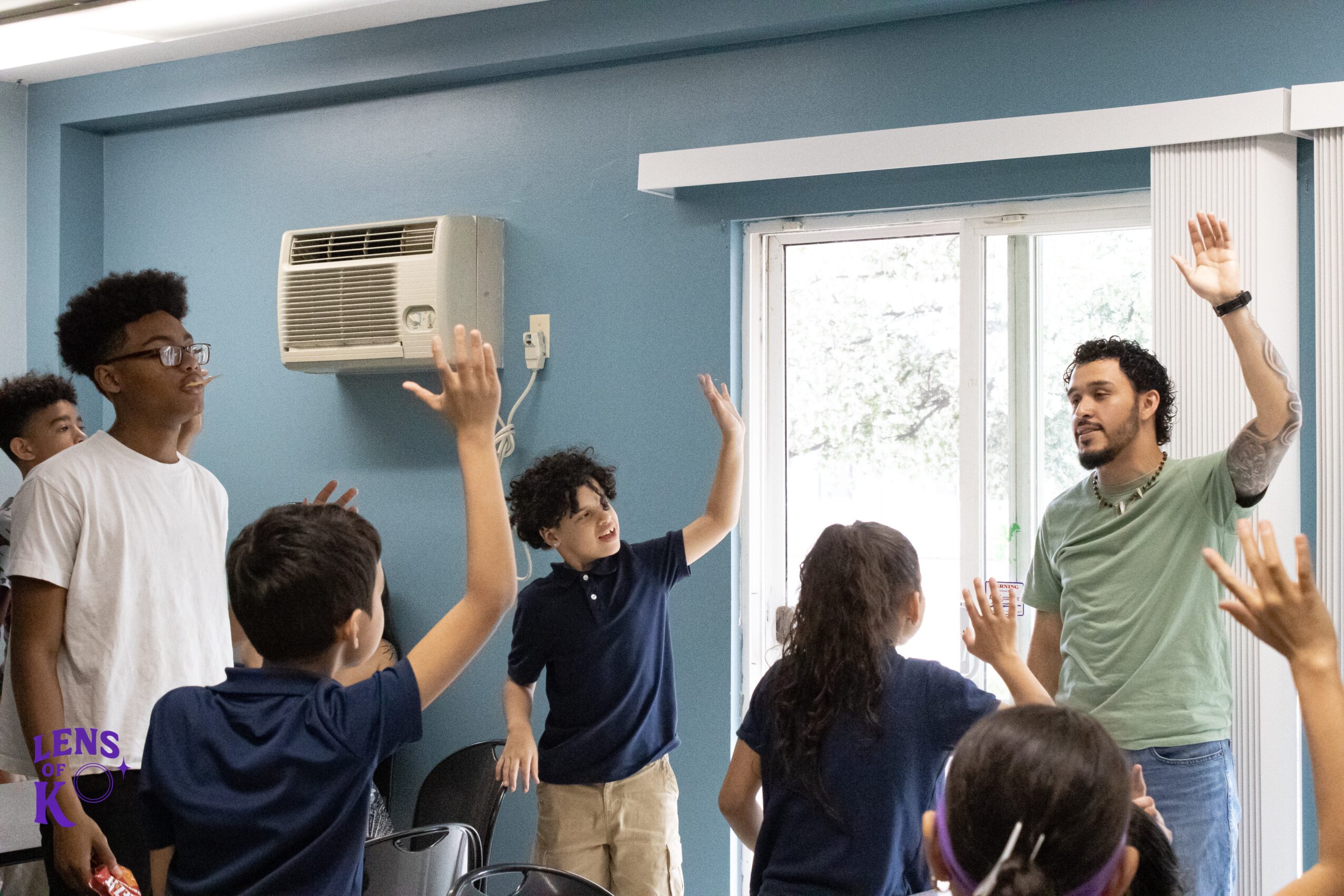 Group of children and an adult in a classroom raising hands, with a window and air conditioner in the background.