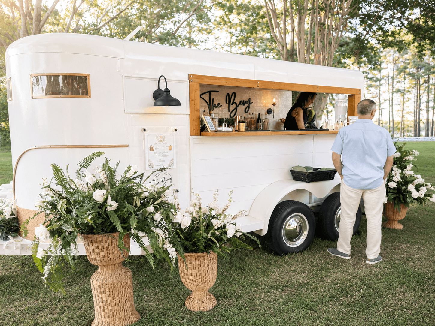 A white mobile bar with a serving window, decorated with large flower arrangements, outdoors with trees in the background.