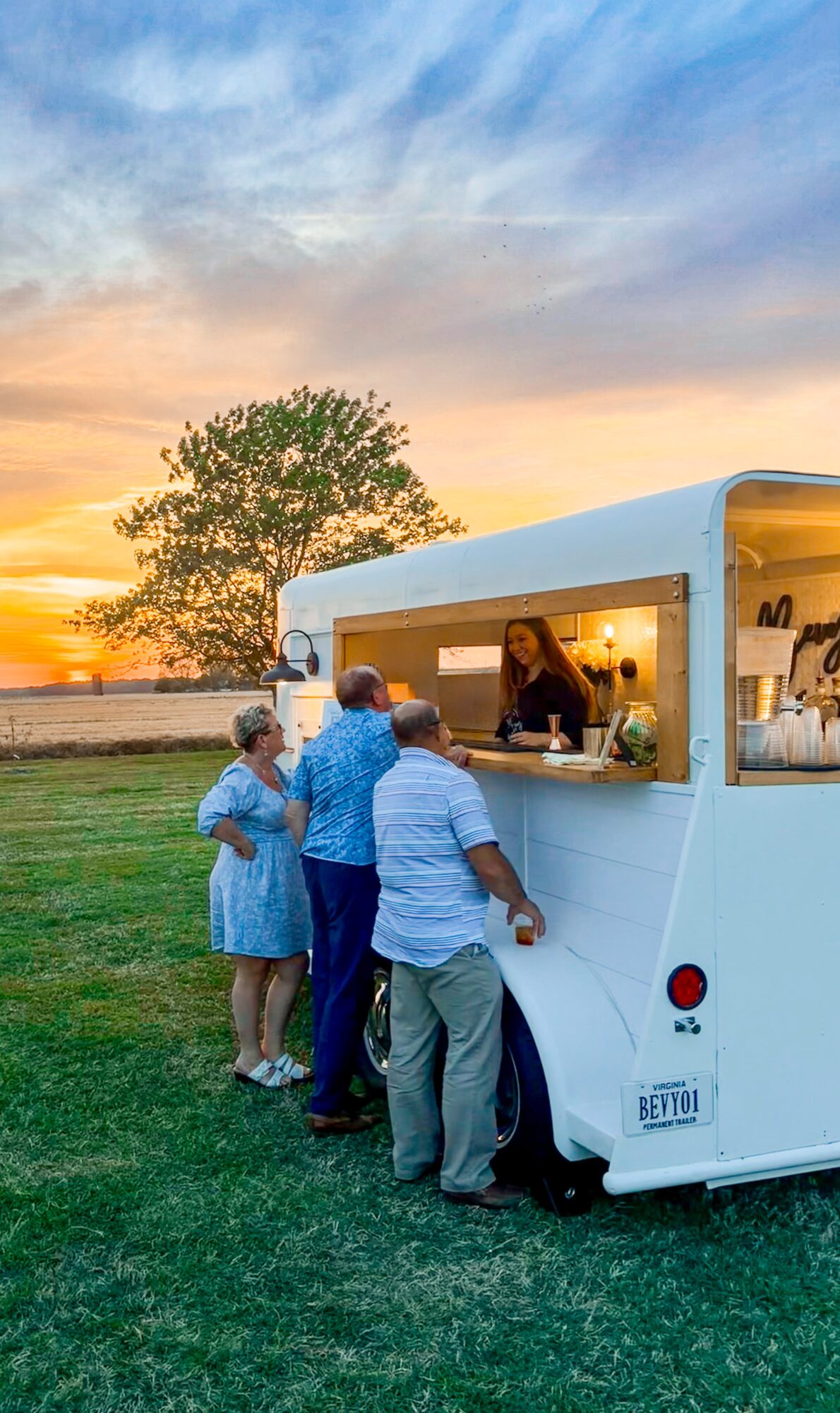 People order food at a mobile food truck during sunset, with a tree and colorful sky in the background.