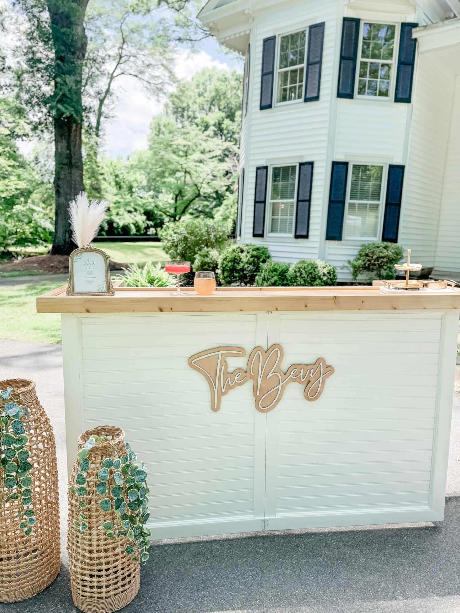 White outdoor bar with a wooden sign reading 'Fitz' in front of a house with blue shutters, surrounded by greenery.