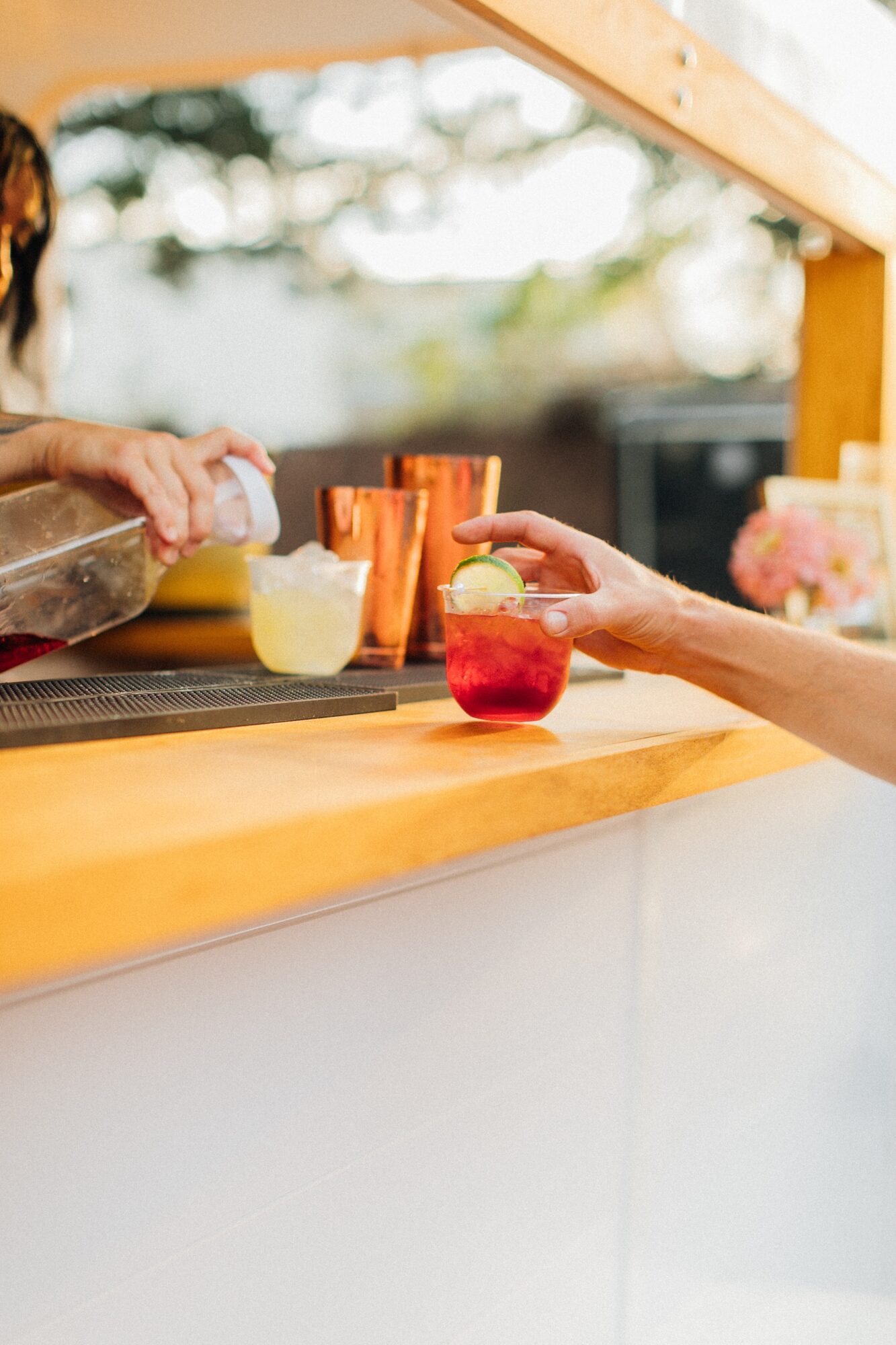 Two people exchanging drinks at a wooden outdoor bar with blurred background, sunlight, and colorful glasses.
