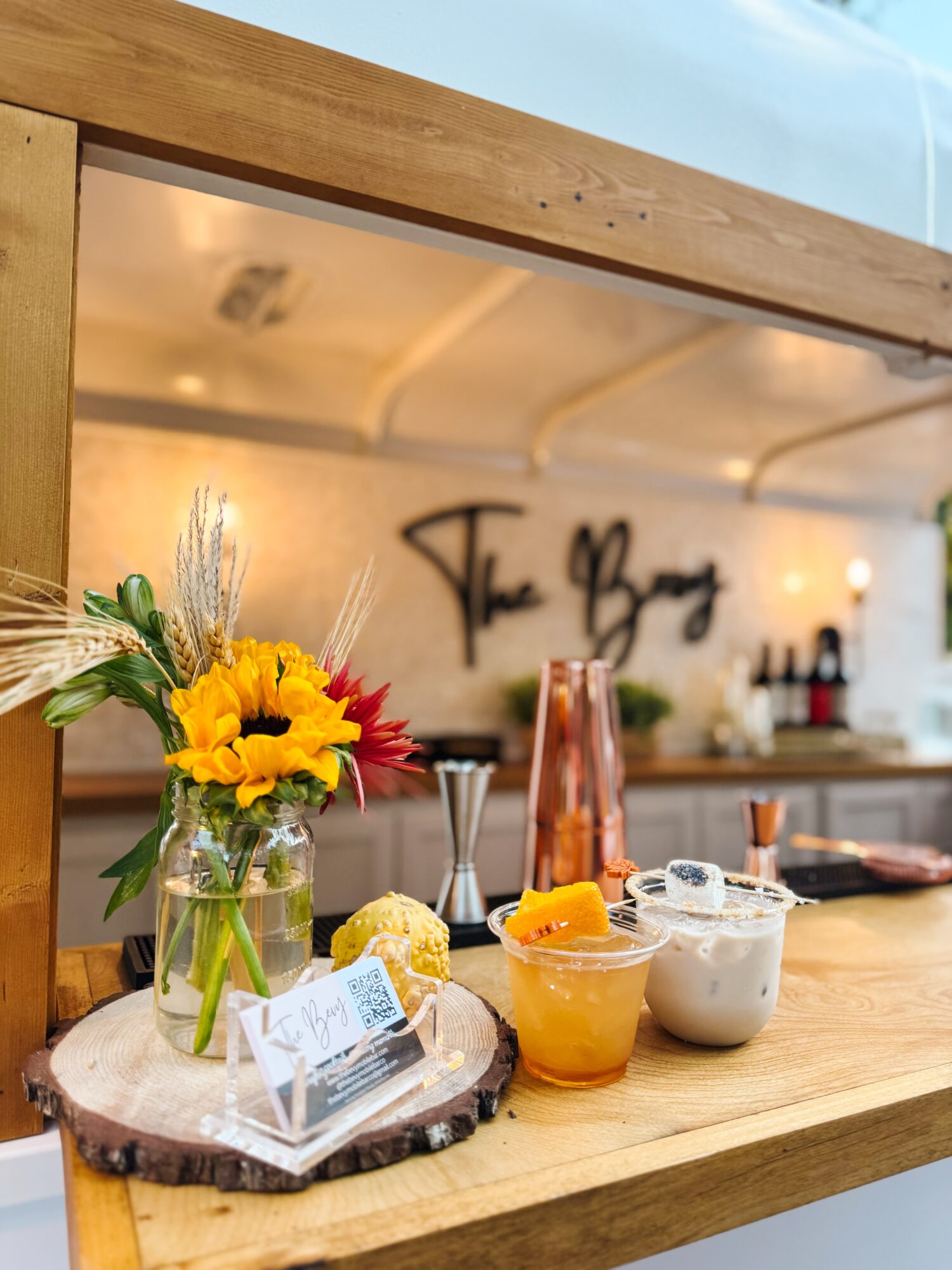 Flower arrangement with sunflowers and greenery, drinks, and a small dessert on a wooden table inside a cafe, with blurred background.