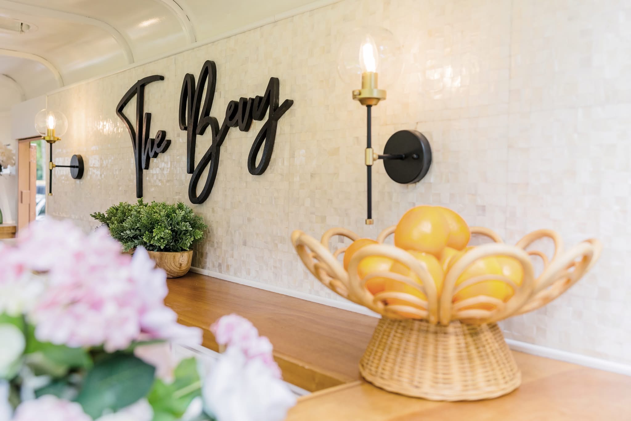 Decorative basket with yellow eggs on a wooden surface, wall with black script sign, wall sconce, and pink flowers in foreground.