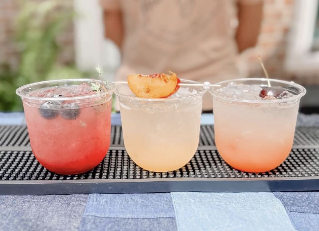 Three colorful cocktails on a black mat, with a person in the background, outdoors, on a sunny day.