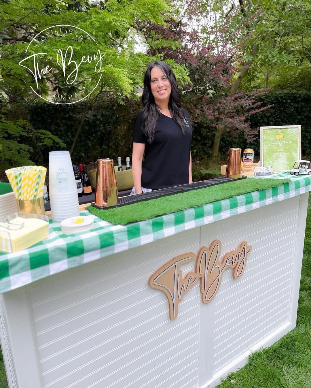 Woman standing behind a beverage stand outdoors with trees in background, green and white checkered tablecloth, cups, and a sign.
