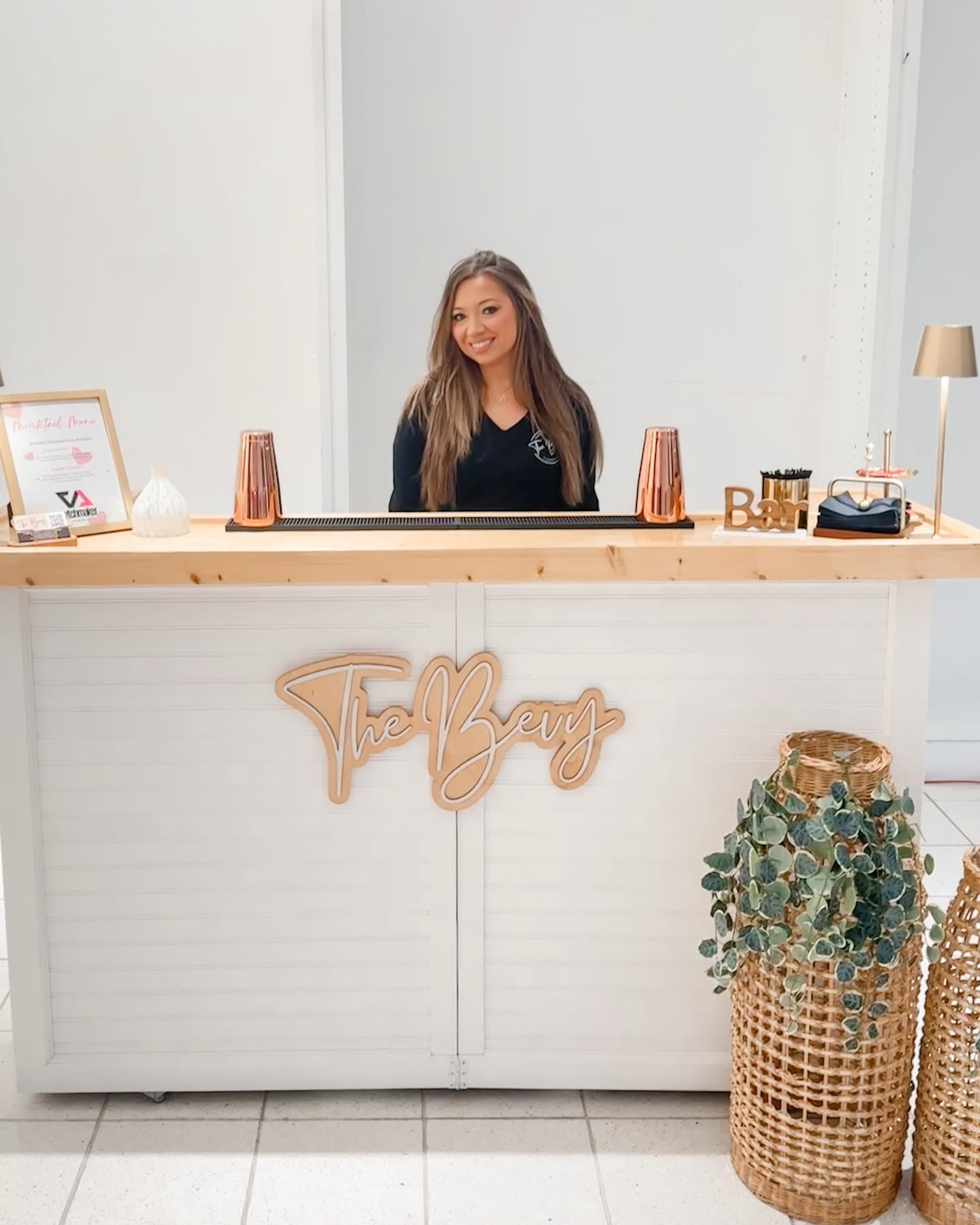 Woman with long hair smiling behind a reception desk with decor and a large basket of plants, in a bright room.