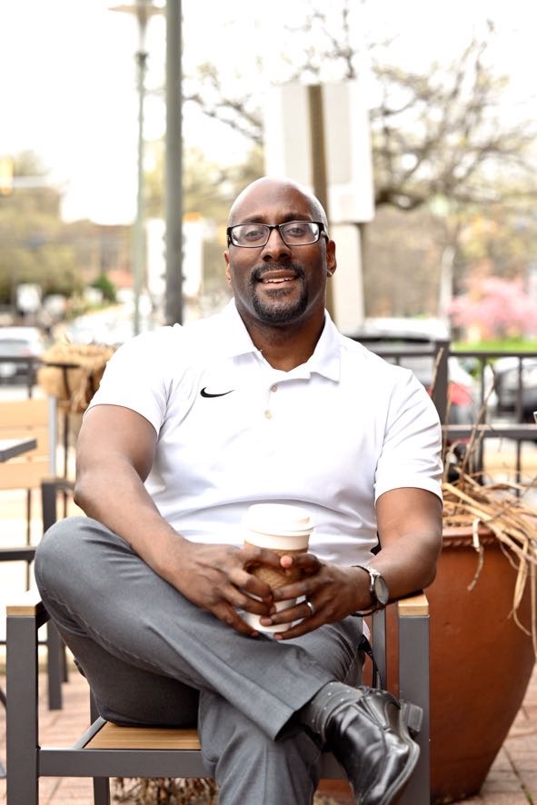 Man sitting outdoors on a chair, holding a drink, smiling, wearing glasses and a white polo shirt.