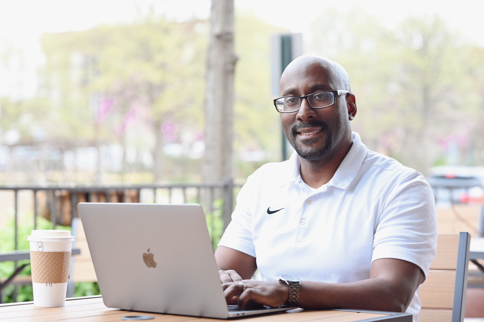Man with glasses and a beard sitting outdoors with a laptop, smiling, holding a coffee cup, trees and a fence in background.