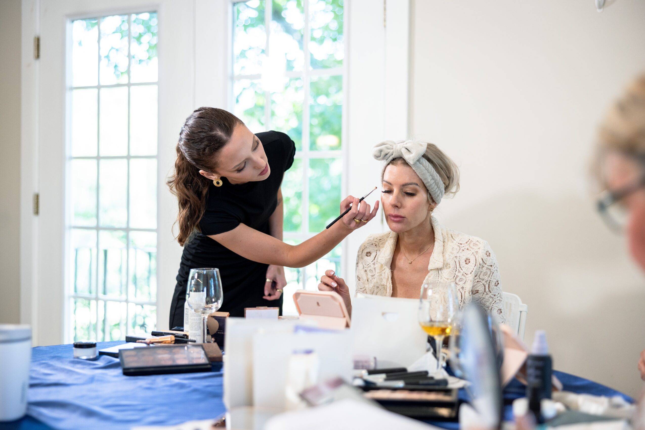 Makeup artist applying makeup to a woman seated at a table with makeup products, in a bright room with large windows.