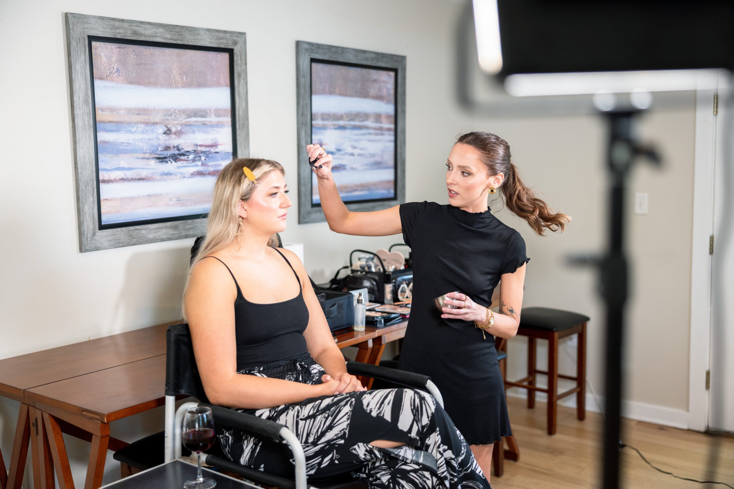 Two women in a room with framed pictures on the wall, one sitting and the other applying makeup or skincare, with a camera in the foreground.