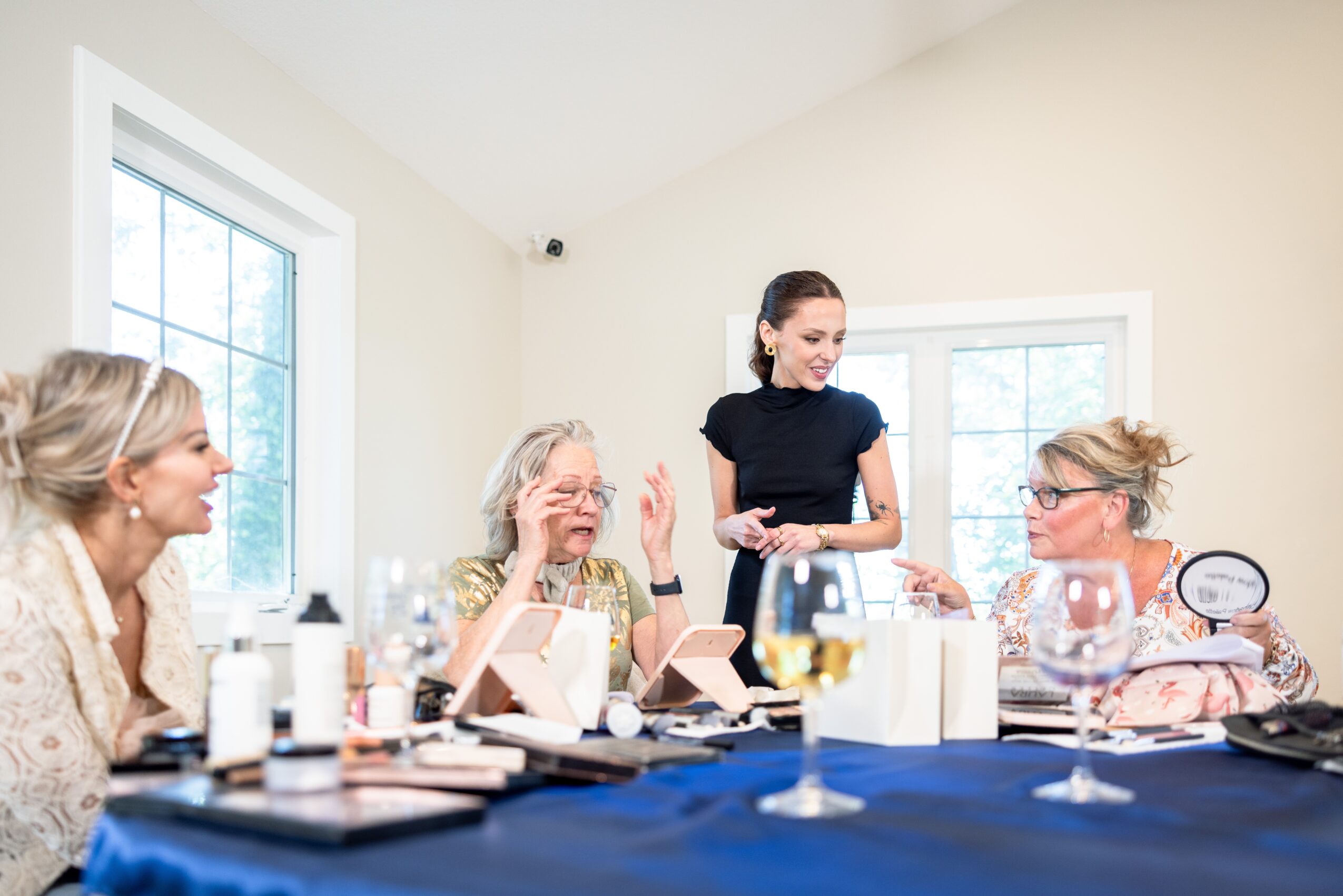 Four women sit around a table with makeup and drinks, engaging in conversation, in a bright room with windows.