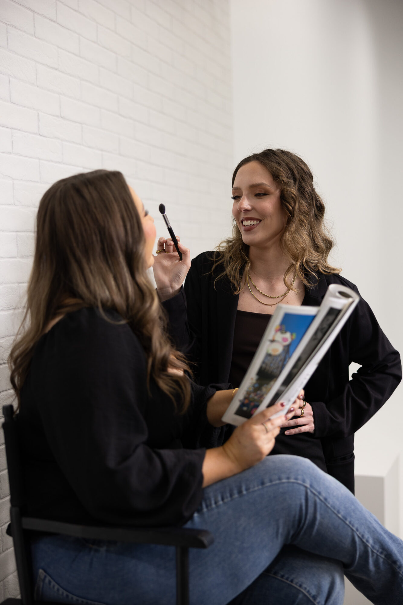 Two women talking, one holding a makeup brush and a magazine, smiling in a bright room with white walls.