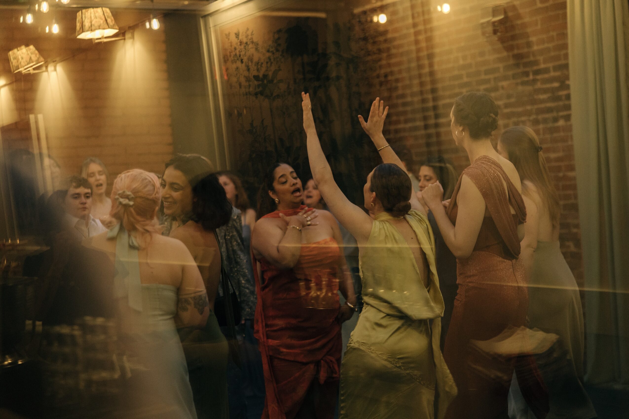 Group of people dancing and socializing indoors with warm lighting and brick walls, some raising hands and talking.