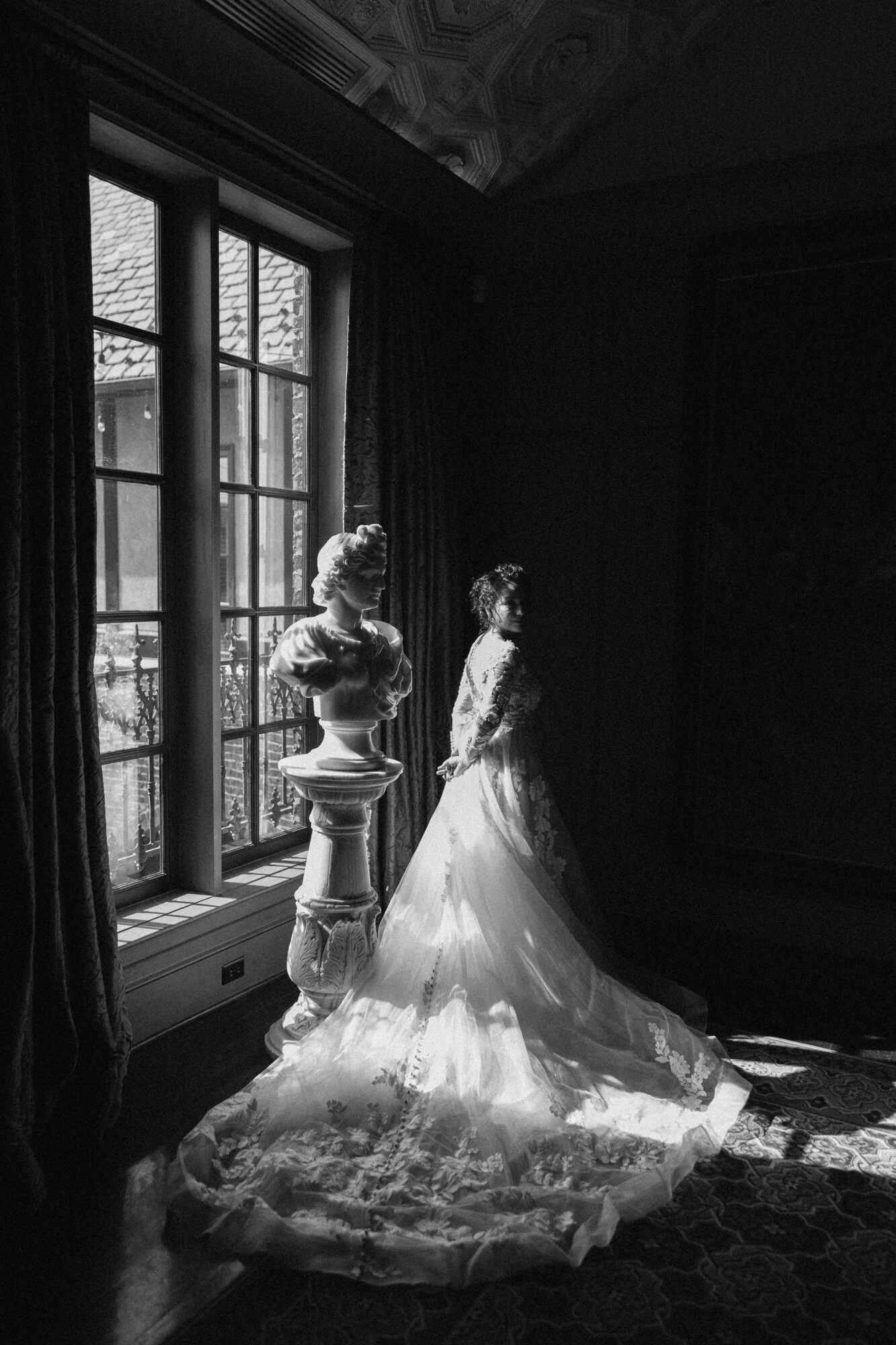 A bride in a wedding dress stands near a large window with sunlight streaming in, casting shadows inside a room.