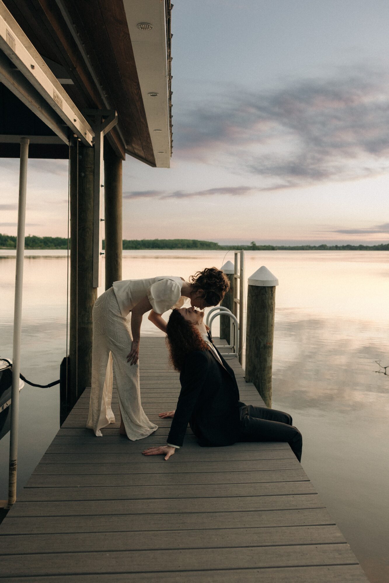Two people on a dock by a calm lake at sunset, one leaning over the other, holding hands, with a building overhead.