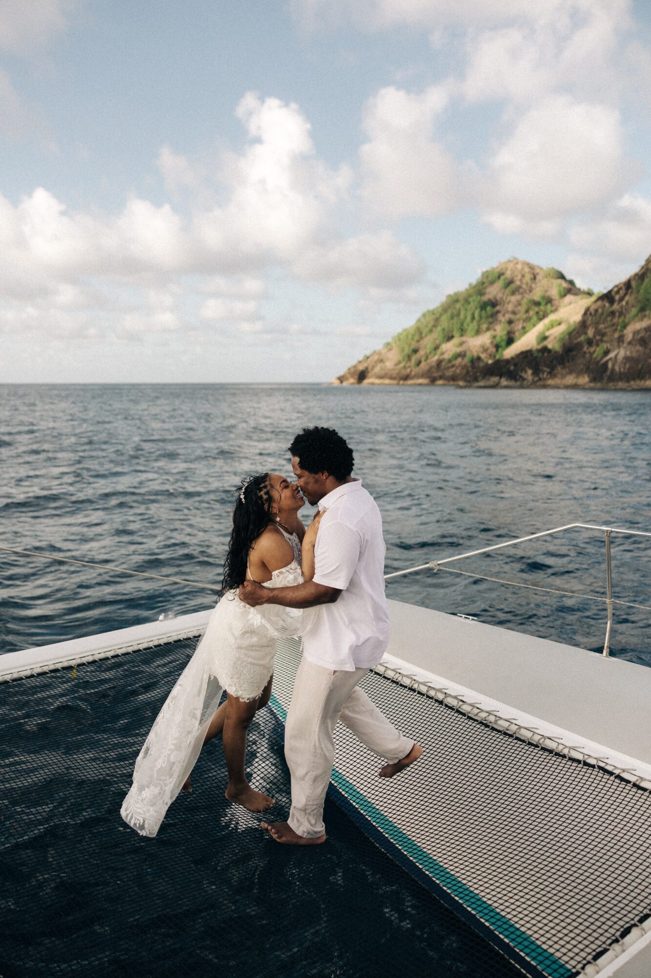 Two people embrace on a boat with a scenic ocean and hillside background, under partly cloudy sky.