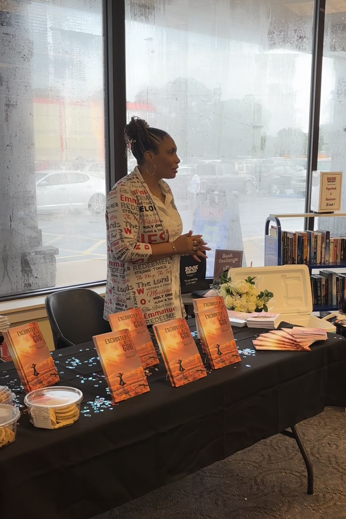Woman standing behind a table with books and promotional materials inside a store, large windows in the background.