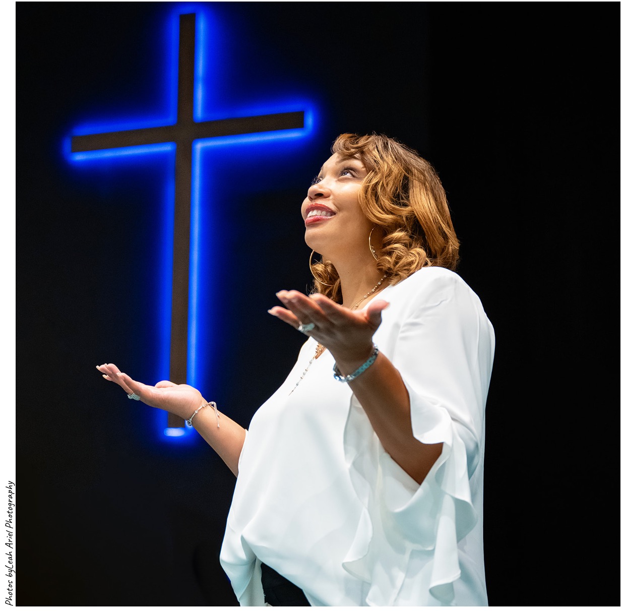 Woman with arms raised, smiling, in front of a glowing blue cross on a dark background.