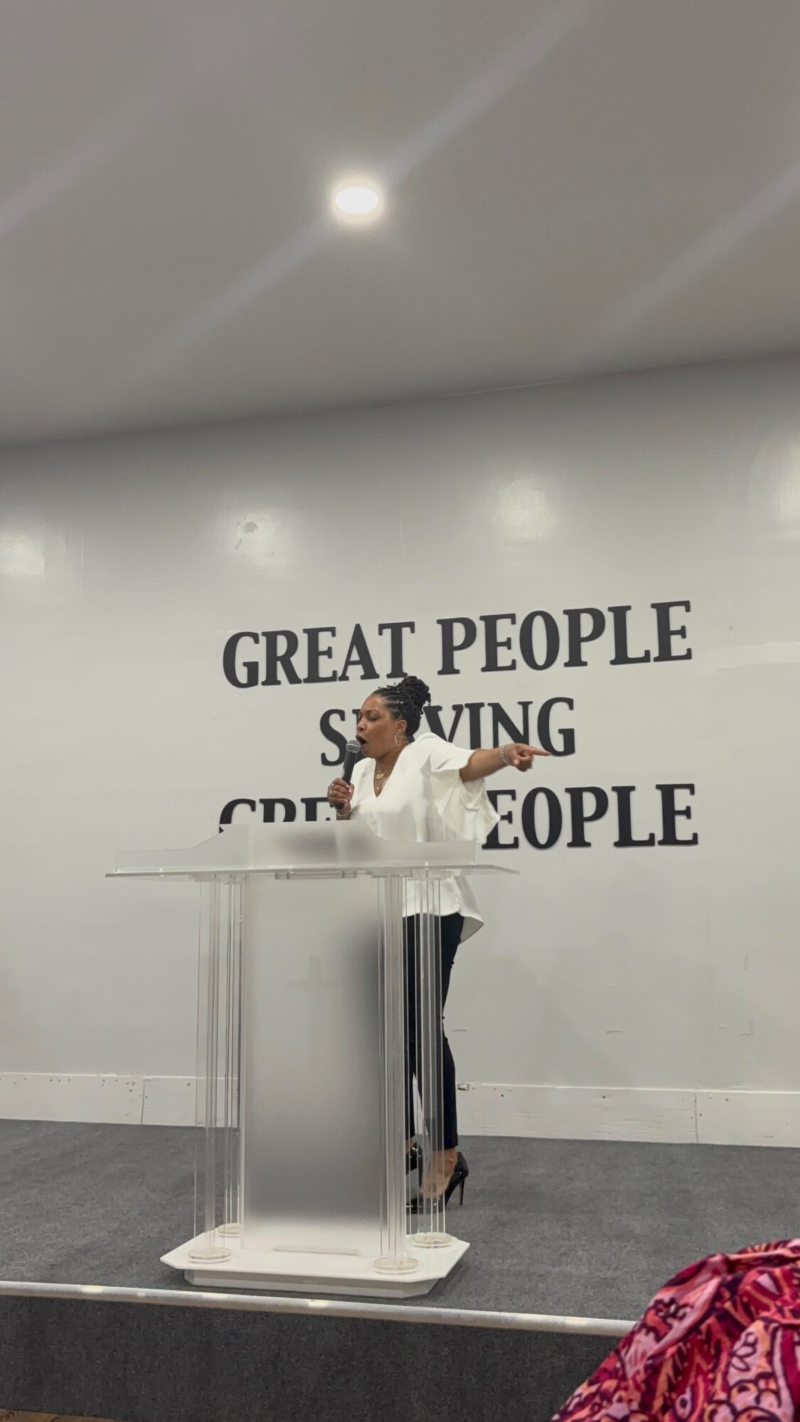 Person speaking at a podium in front of a wall with the words 'GREAT PEOPLE SAVING GREAT PEOPLE'.