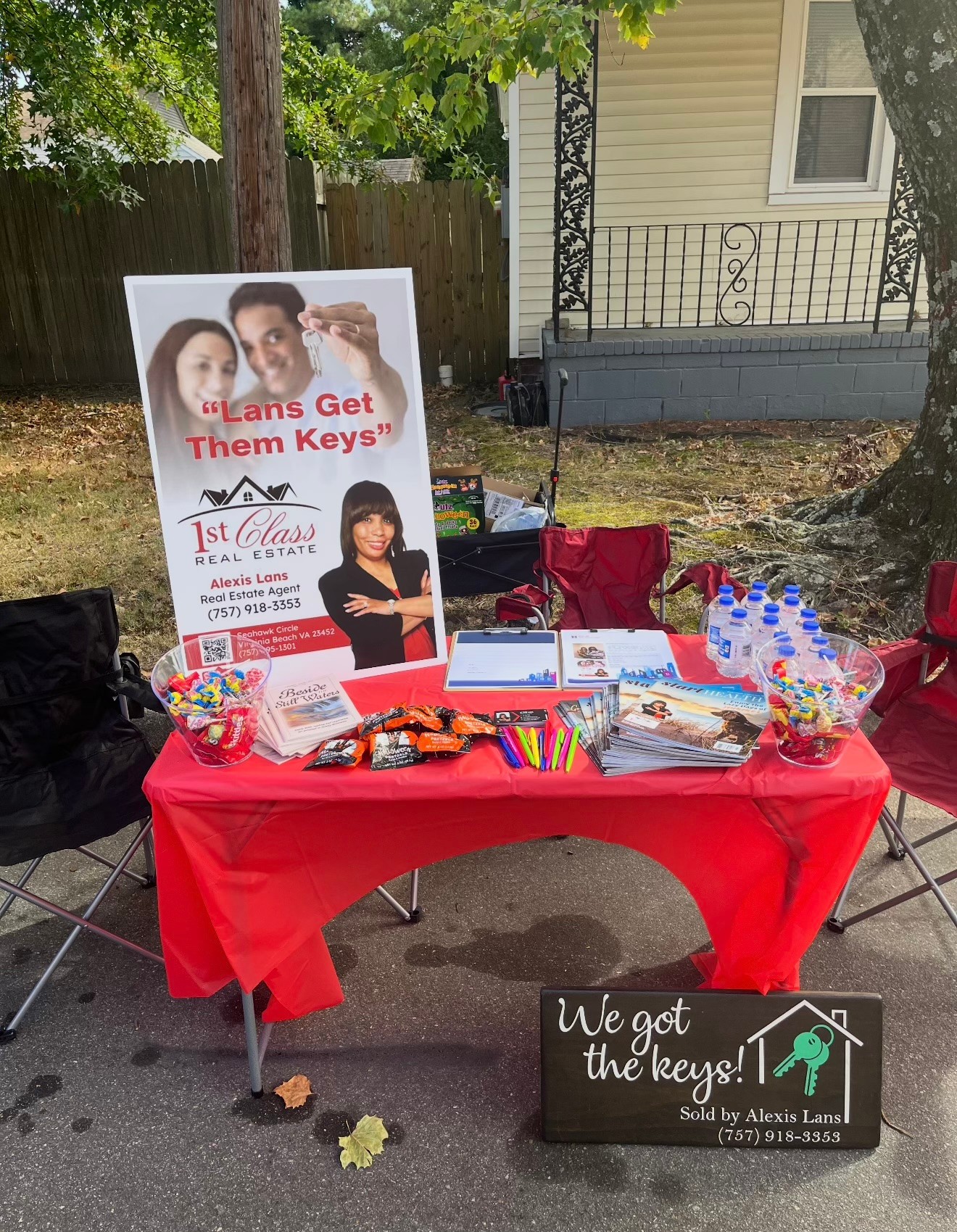 Table with red tablecloth displaying books, pens, and promotional materials outdoors, with a sign about unlocking keys and a large poster featuring two women and text about earning keys, in a yard with trees and a house in the background.