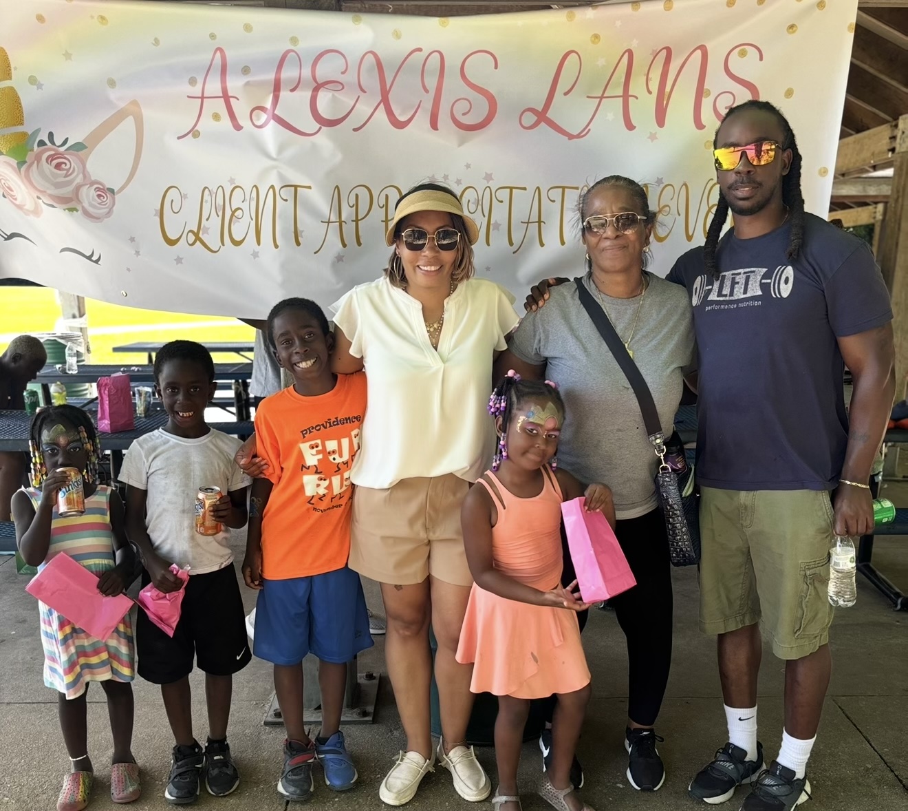 Group of people standing in front of a banner, smiling, with children and adults, at an outdoor event.