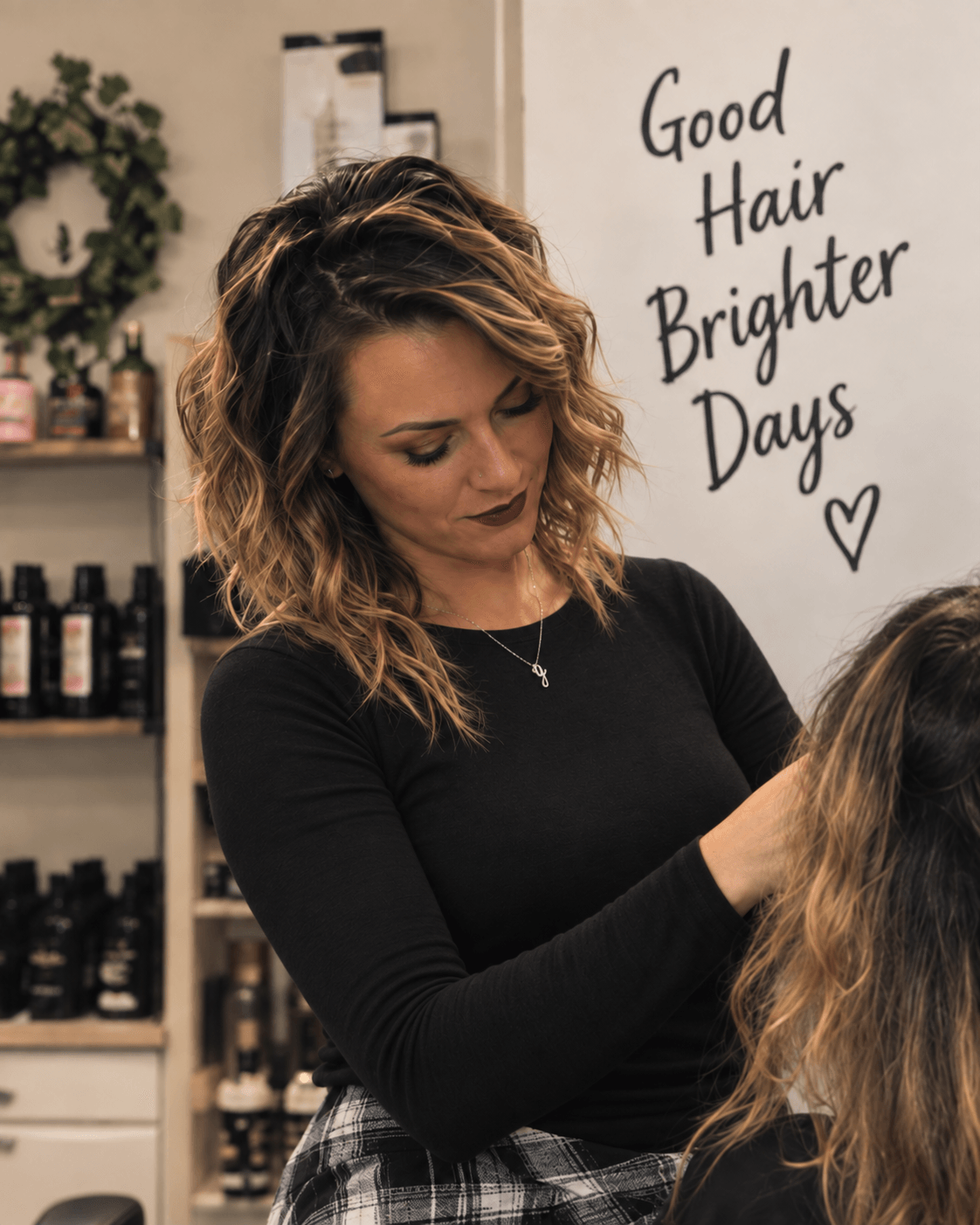 Woman with wavy hair in a salon, looking down, with shelves of hair products behind her, and a sign reading 'Good Hair Brighter Days' with a heart.