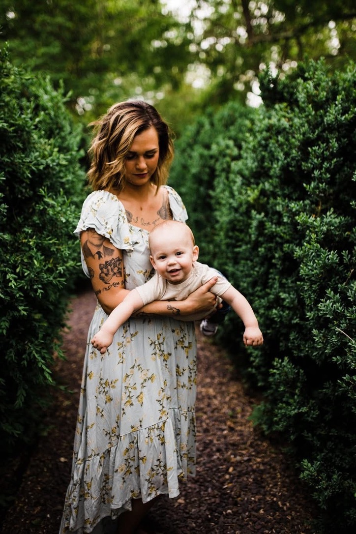 Woman holding a smiling baby outdoors among green bushes, with trees in the background.