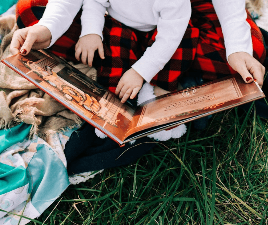 Two children sitting on grass, reading a large illustrated book, with a blanket and bag nearby.