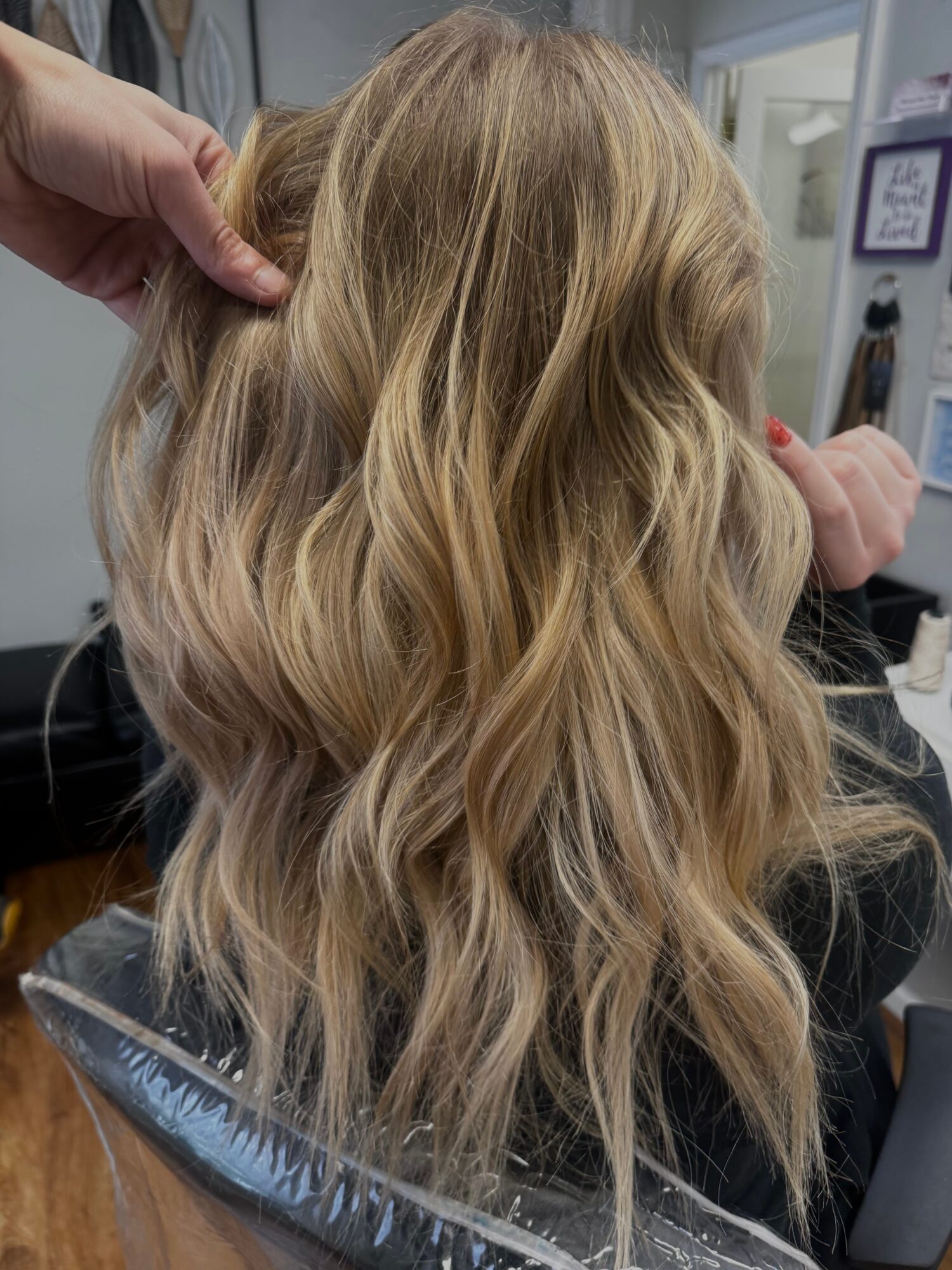 Person with long, wavy blonde hair, viewed from behind, in a salon setting.