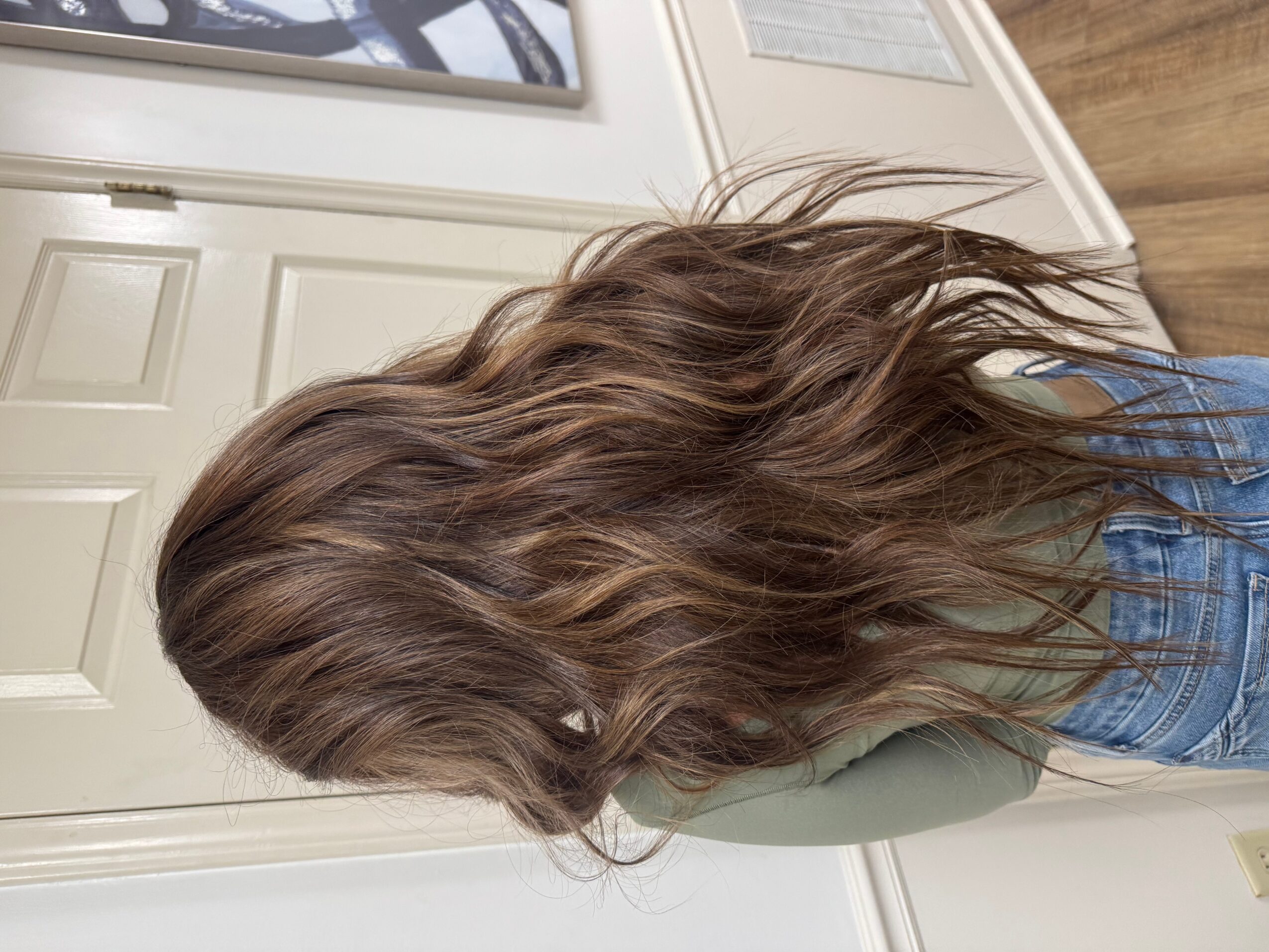 Side view of a person with long, wavy brown hair, wearing a blue shirt, indoors near a white door.