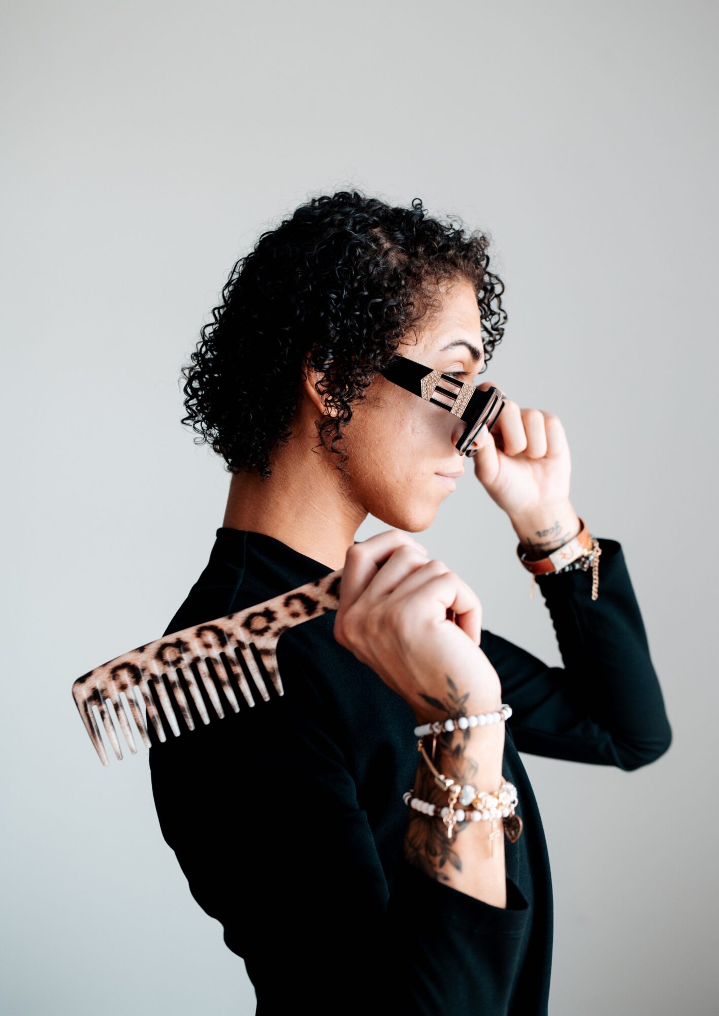Woman with curly hair holding a comb and a hair clip, wearing bracelets and a black top, against a plain background.