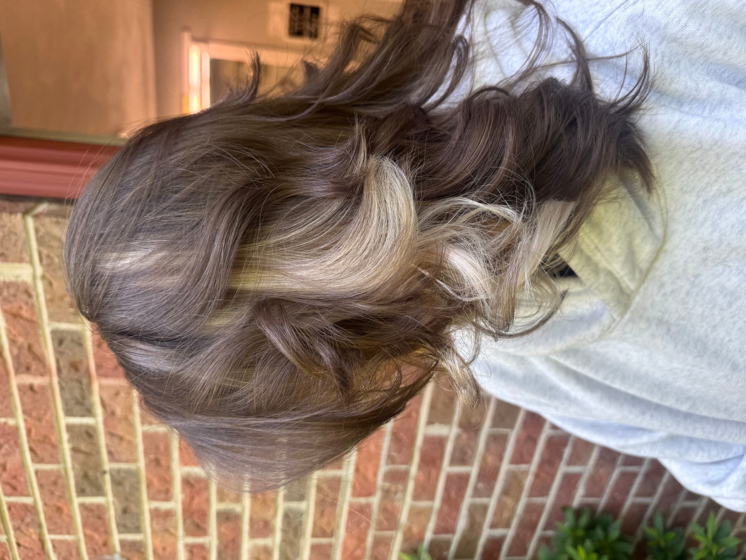Person with long, wavy hair, light and dark shades, standing outdoors near a brick wall.