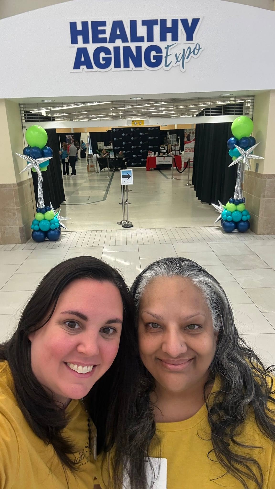 Two women in yellow shirts smiling in front of a sign that reads 'Healthy Aging Expo' at an indoor event.