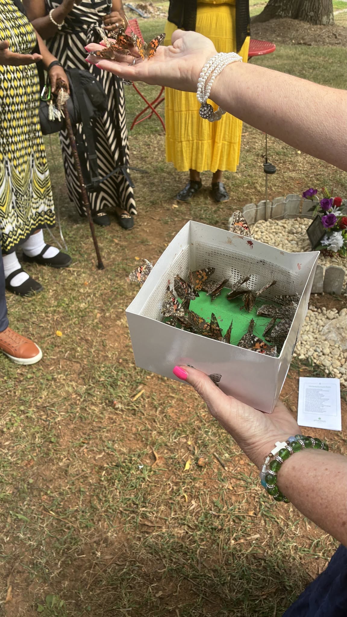Person holding a box with a green object inside, surrounded by people outdoors, some wearing yellow and patterned clothing.