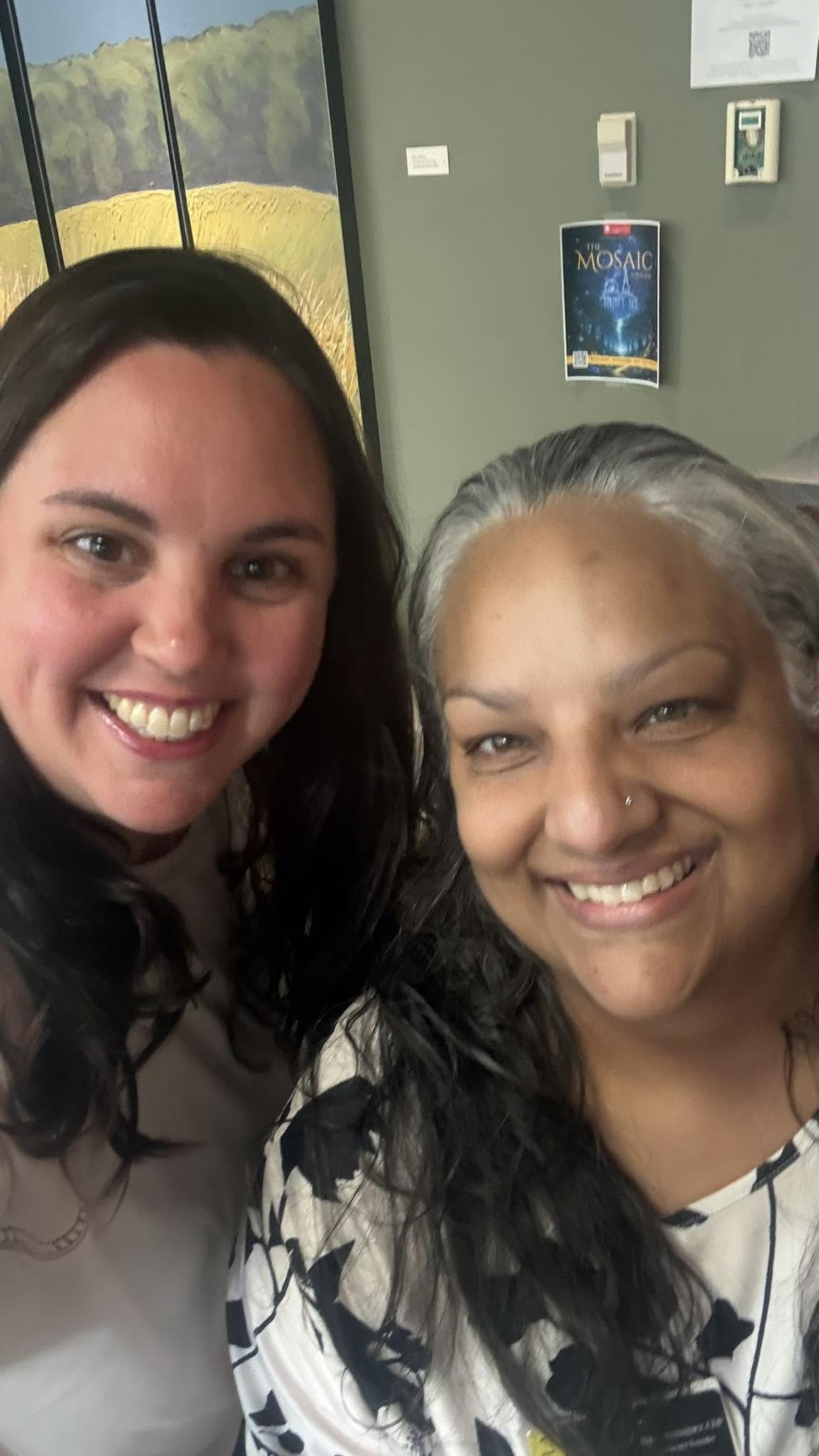 Two women smiling indoors, one with dark hair and the other with gray hair, background includes a painting and a poster.