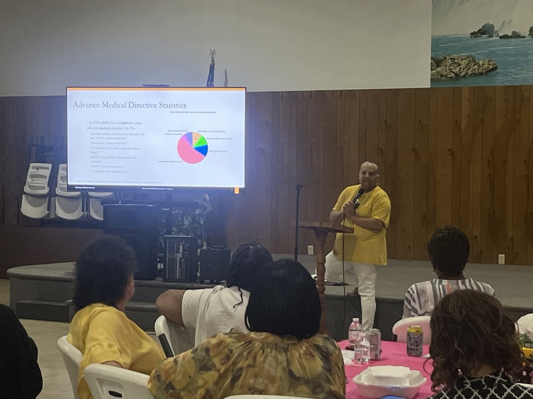 A man in a yellow shirt stands at a podium giving a presentation to an audience in a room with wooden walls and a large screen displaying a slide with a pie chart.