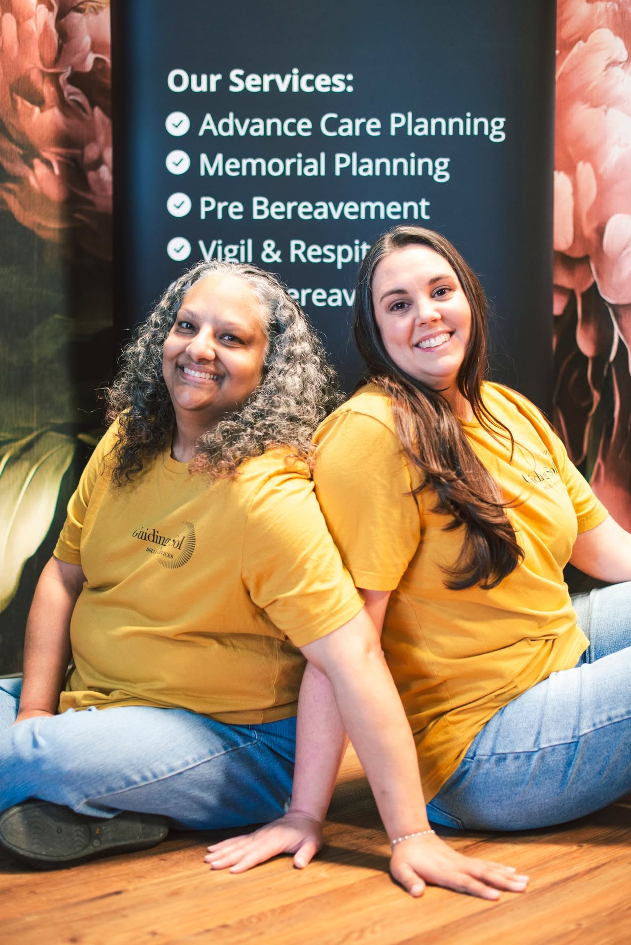 Two women sitting on the floor in front of a blue sign with service list, smiling, wearing yellow shirts, with floral background.