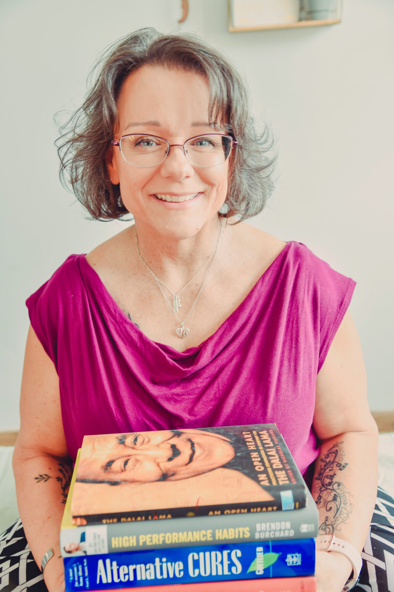 Smiling woman with glasses and short gray hair holding a book, wearing a pink top, in a room with light-colored walls.
