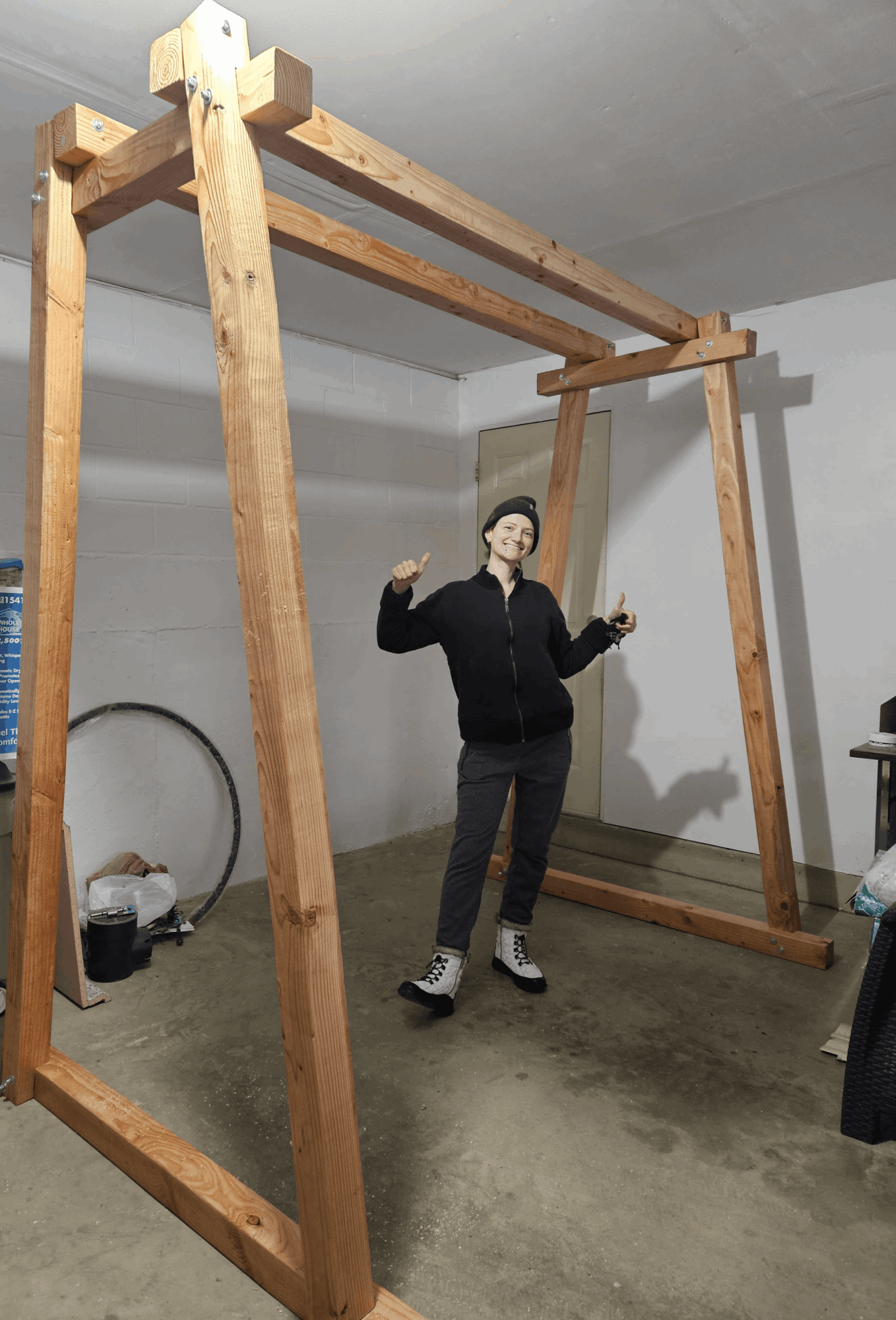 Person standing inside a large wooden frame structure, smiling and making a peace sign with both hands, in a room with concrete floor.