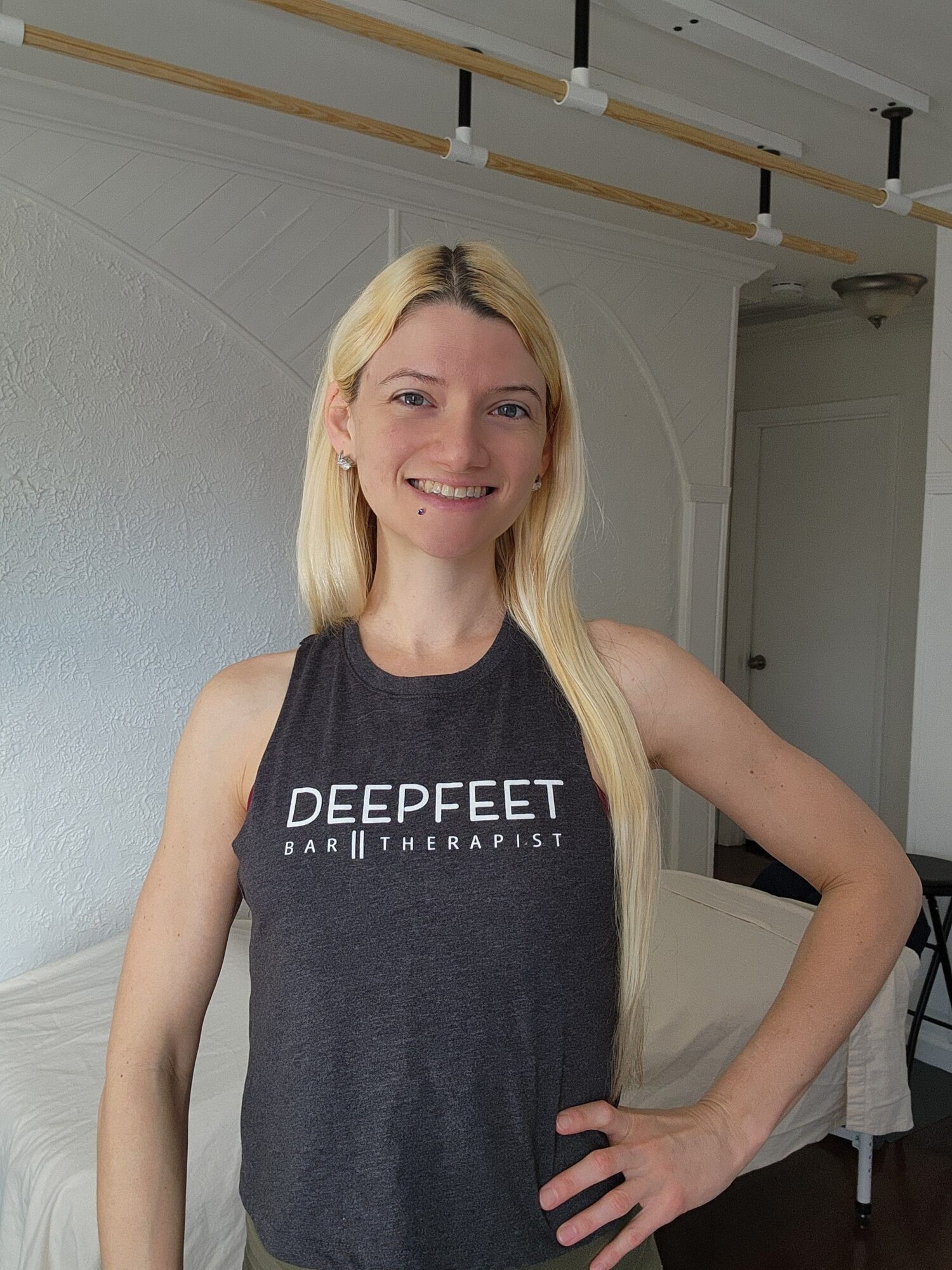 Woman with long blonde hair wearing a black sleeveless shirt, smiling, standing indoors against a white wall.