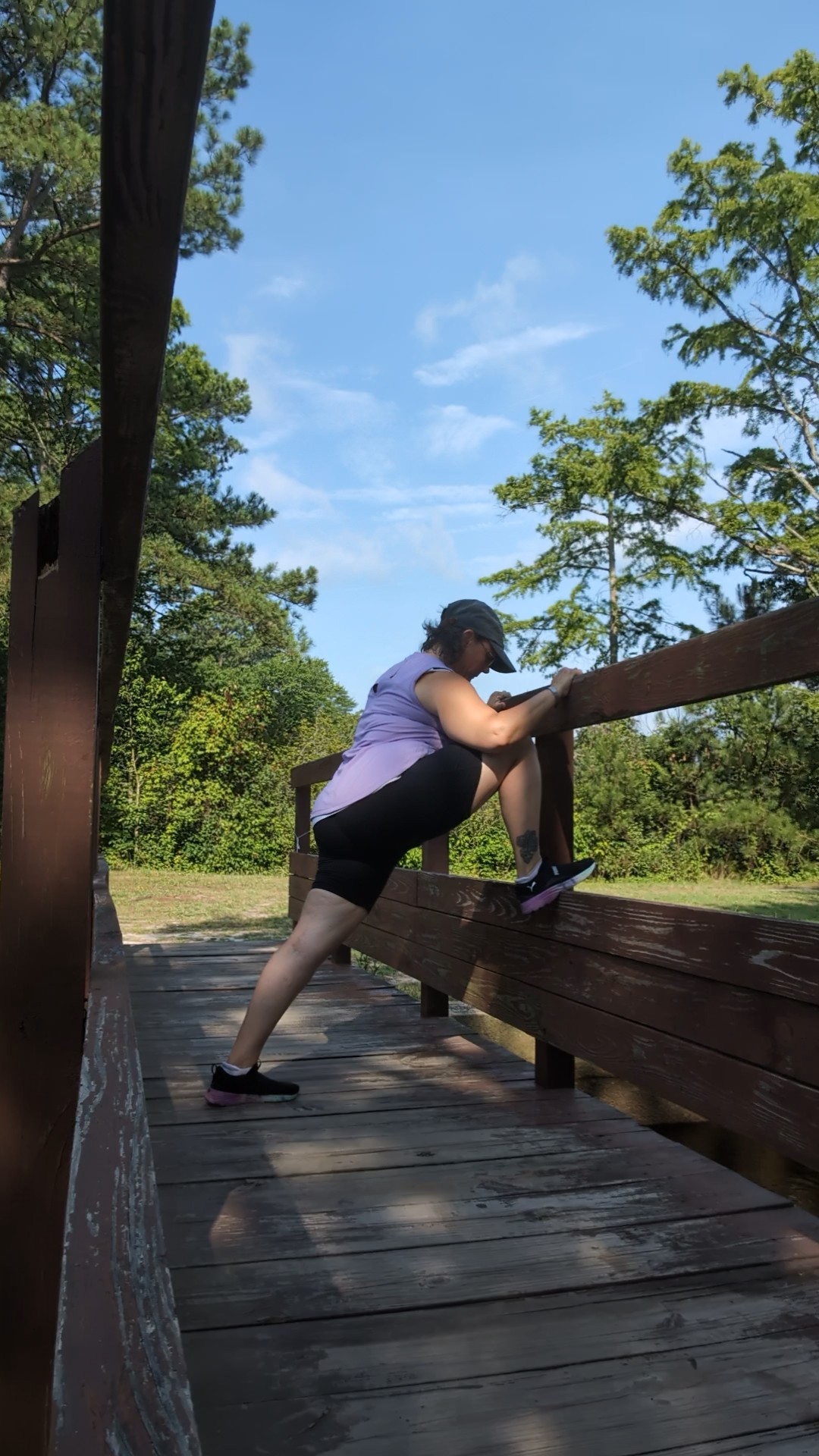 Person stretching on a wooden bridge outdoors with trees and blue sky in background.