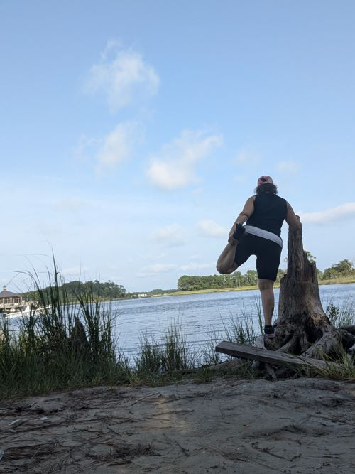 Person standing on a tree stump by a river, looking at the water, with a blue sky and clouds above.
