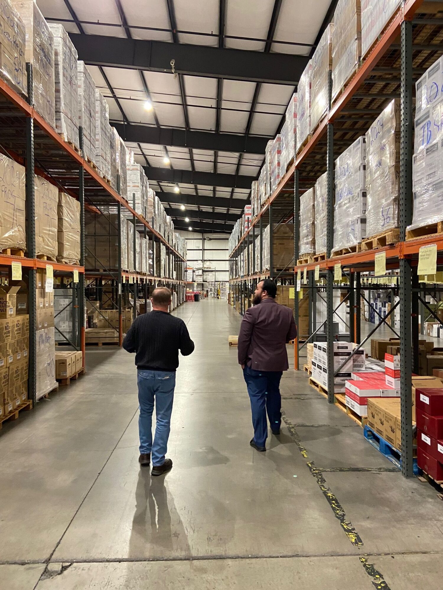 Two people walk through a warehouse aisle with shelves stocked with boxes and products, concrete floor, industrial setting.